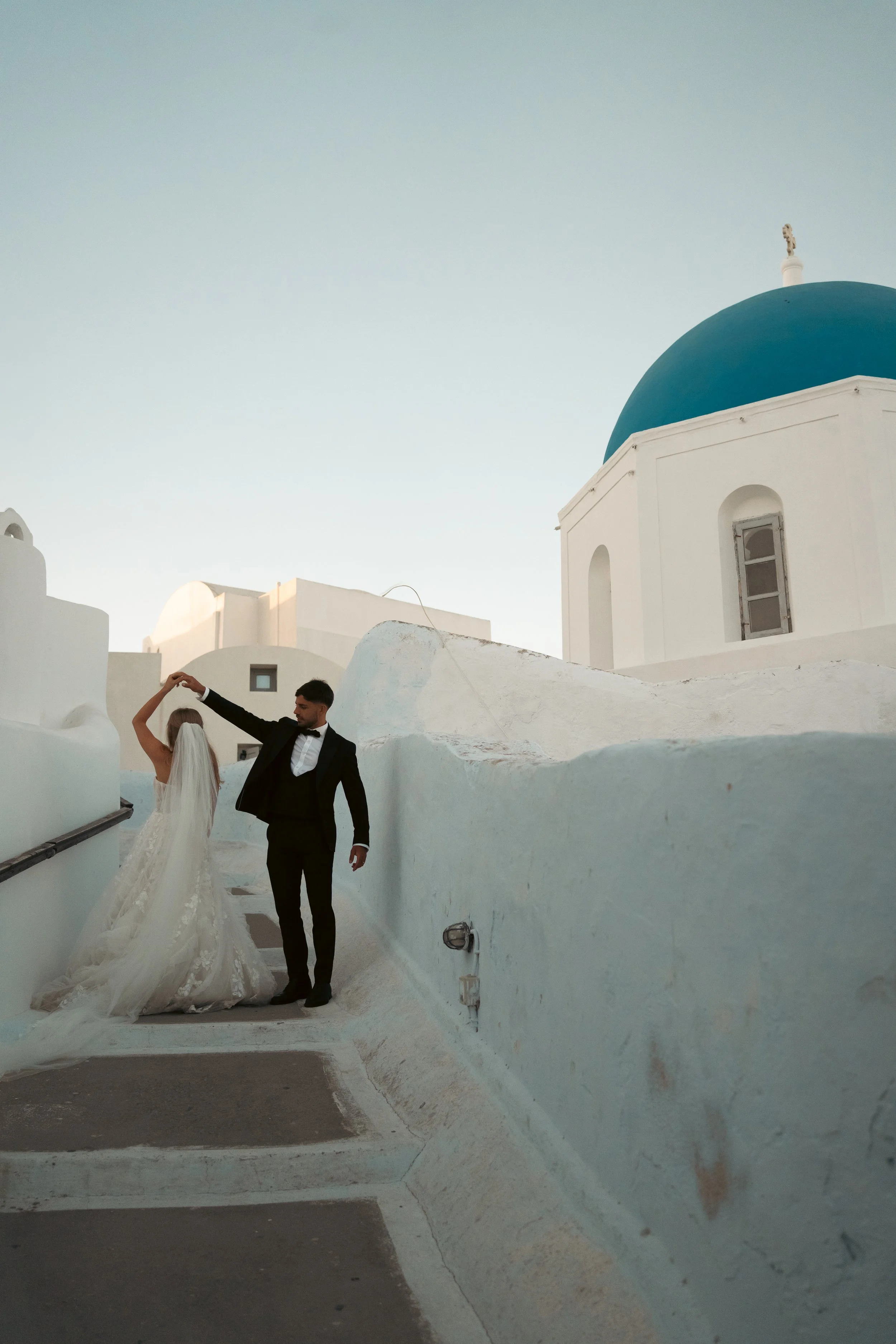 A bride and groom dancing on a white staircase outside a white building with a blue domed roof, typical of Greek architecture in Santorini.