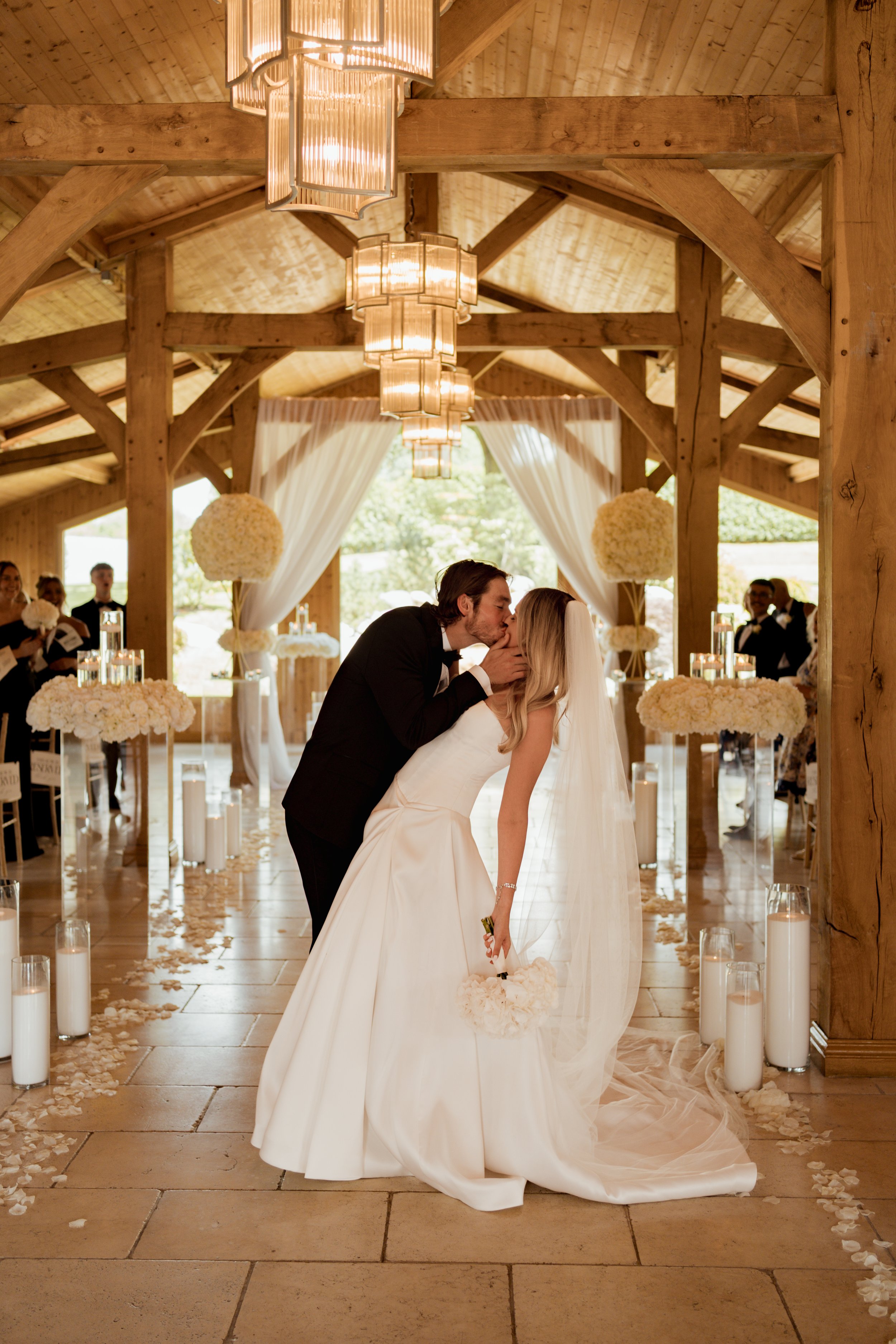 A bride and groom share a kiss during their wedding ceremony in a wooden indoor venue decorated with white flowers and candles. The bride wears a white gown and veil, holding a bouquet, while the groom is in a black tuxedo. Guests are seen in the bac