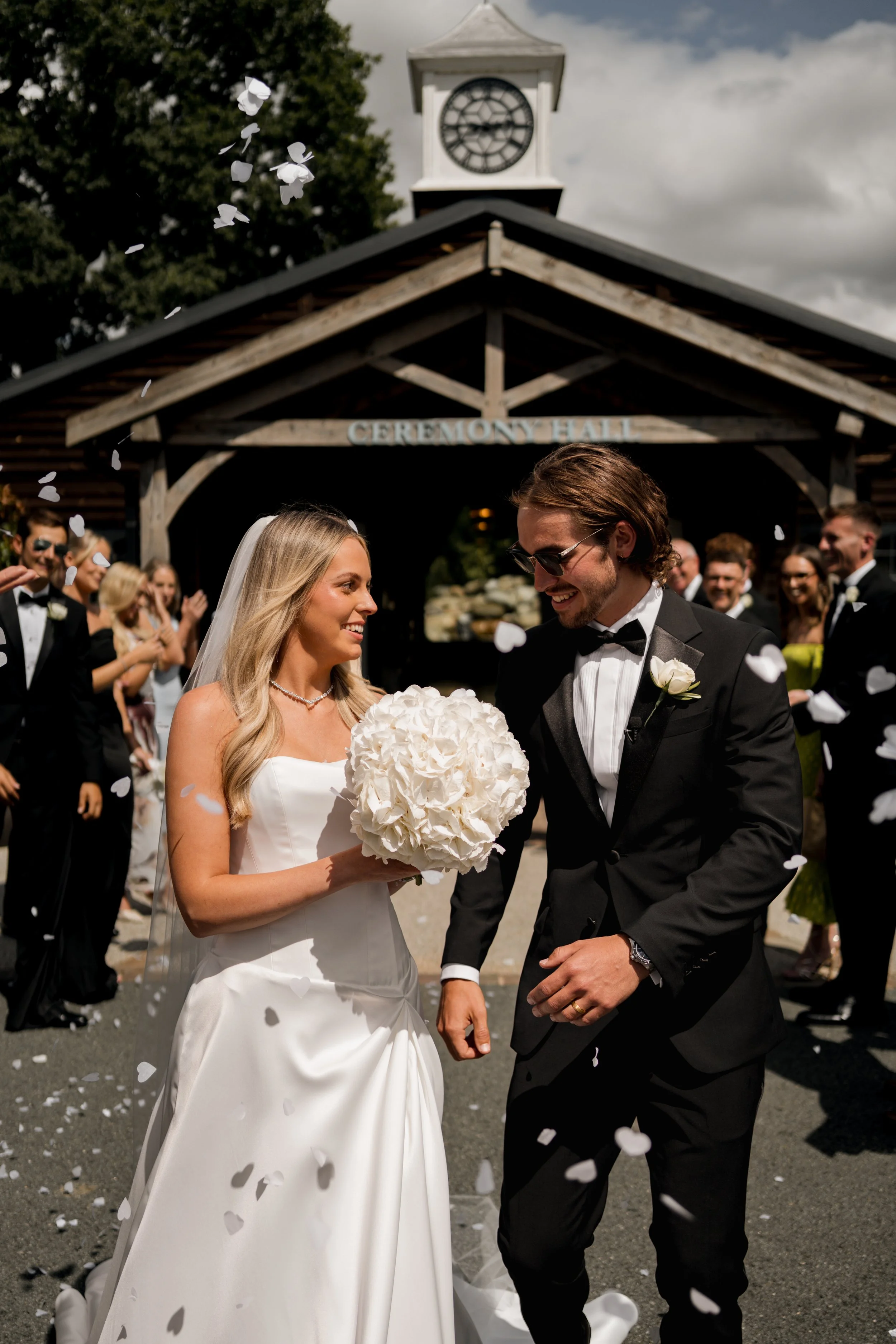 A wedding scene with a bride in a white dress holding a bouquet of white flowers and a groom in a black tuxedo. They are smiling and walking under a wooden ceremony hall with guests celebrating in the background. Confetti is falling around them.