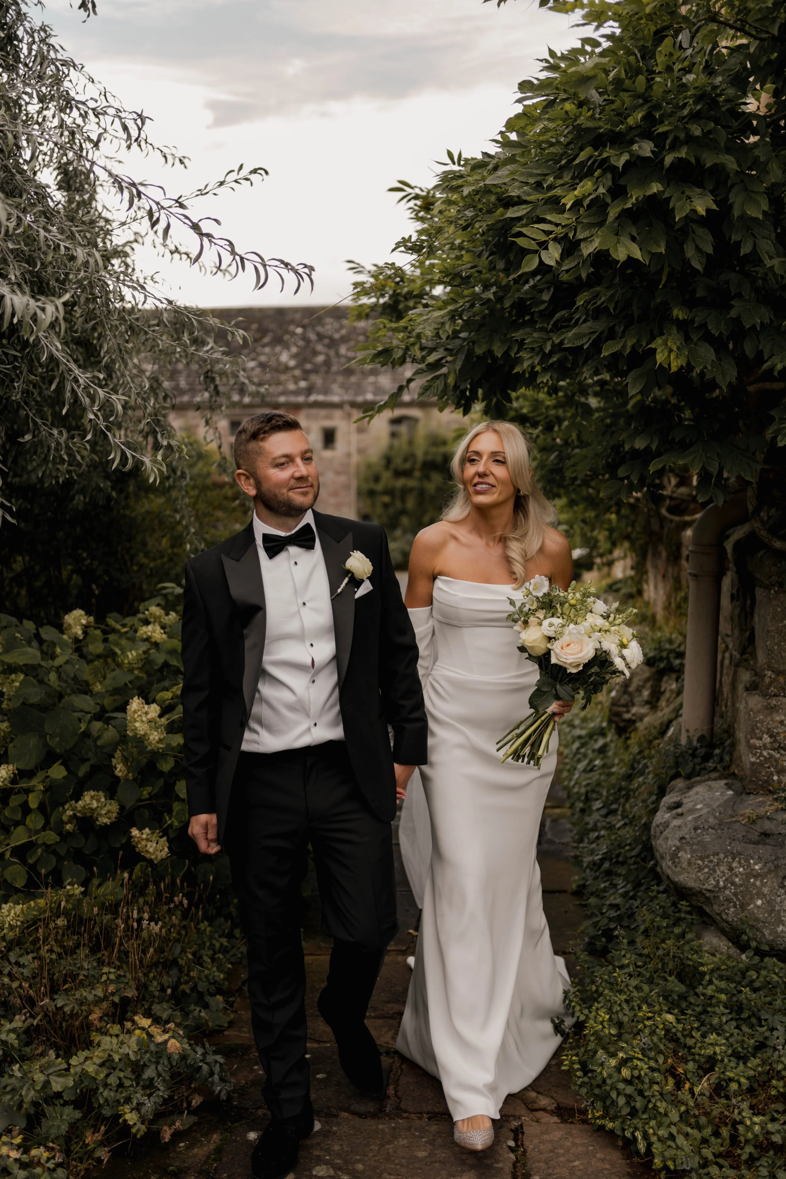 A newlywed couple walking outdoors on a garden pathway, with the bride holding a bouquet of white and pink flowers, smiling, and the groom dressed in a black tuxedo with a bow tie, both surrounded by lush greenery at Askham Hall