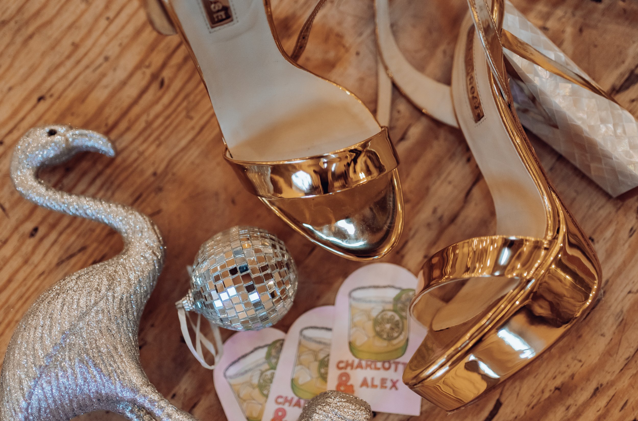 Gold high-heeled shoes, a silver glittery swan figurine, a small disco ball, and pink coasters with a drink illustration and the names Charlotte & Alex on a wooden surface.