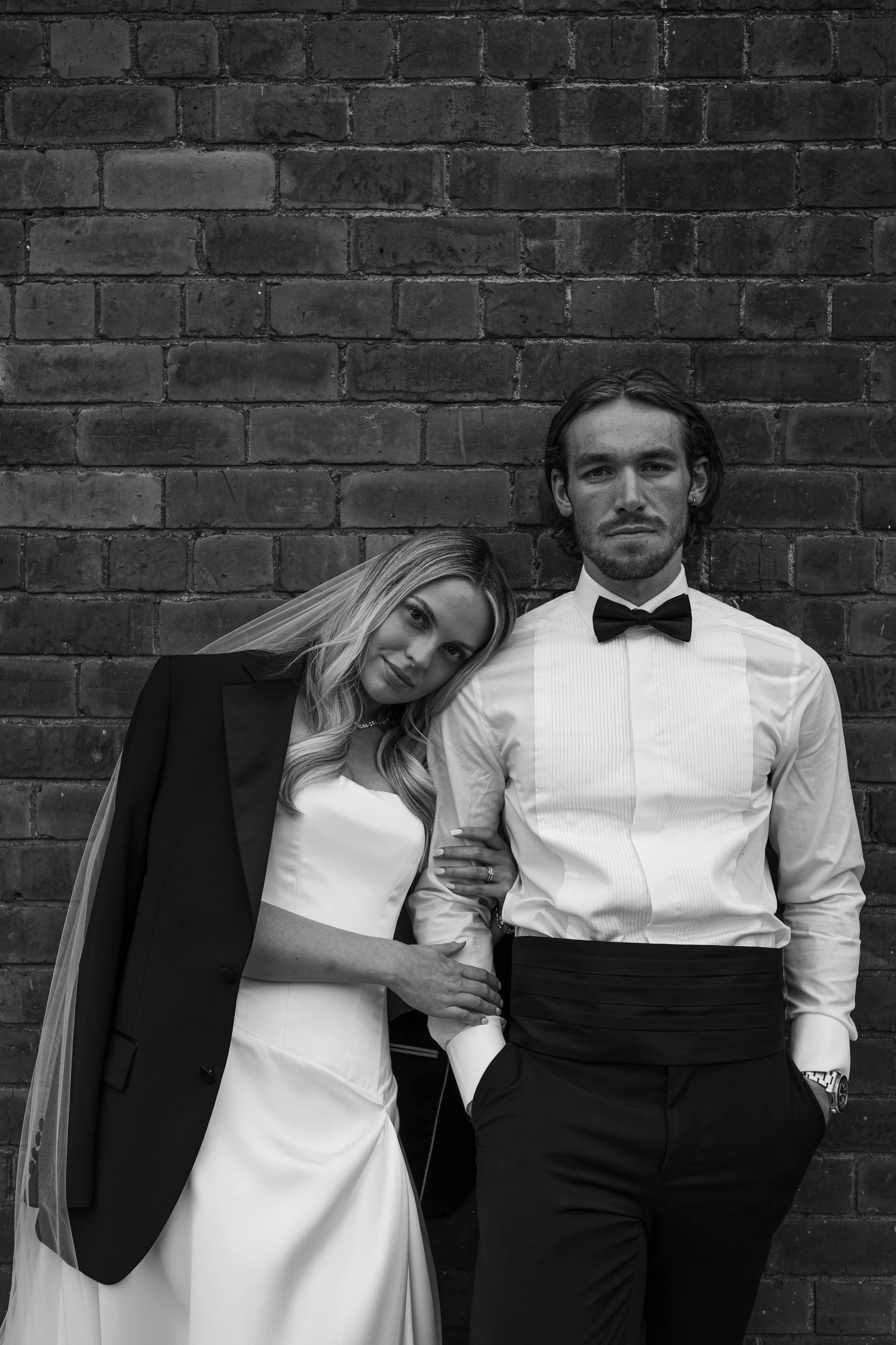 A black-and-white photo of a bride and groom standing against a brick wall. The bride is wearing a veil, a satin wedding dress, and a blazer draped over her shoulders. The groom is dressed in a tuxedo shirt with a bow tie, black pants, and has his ha
