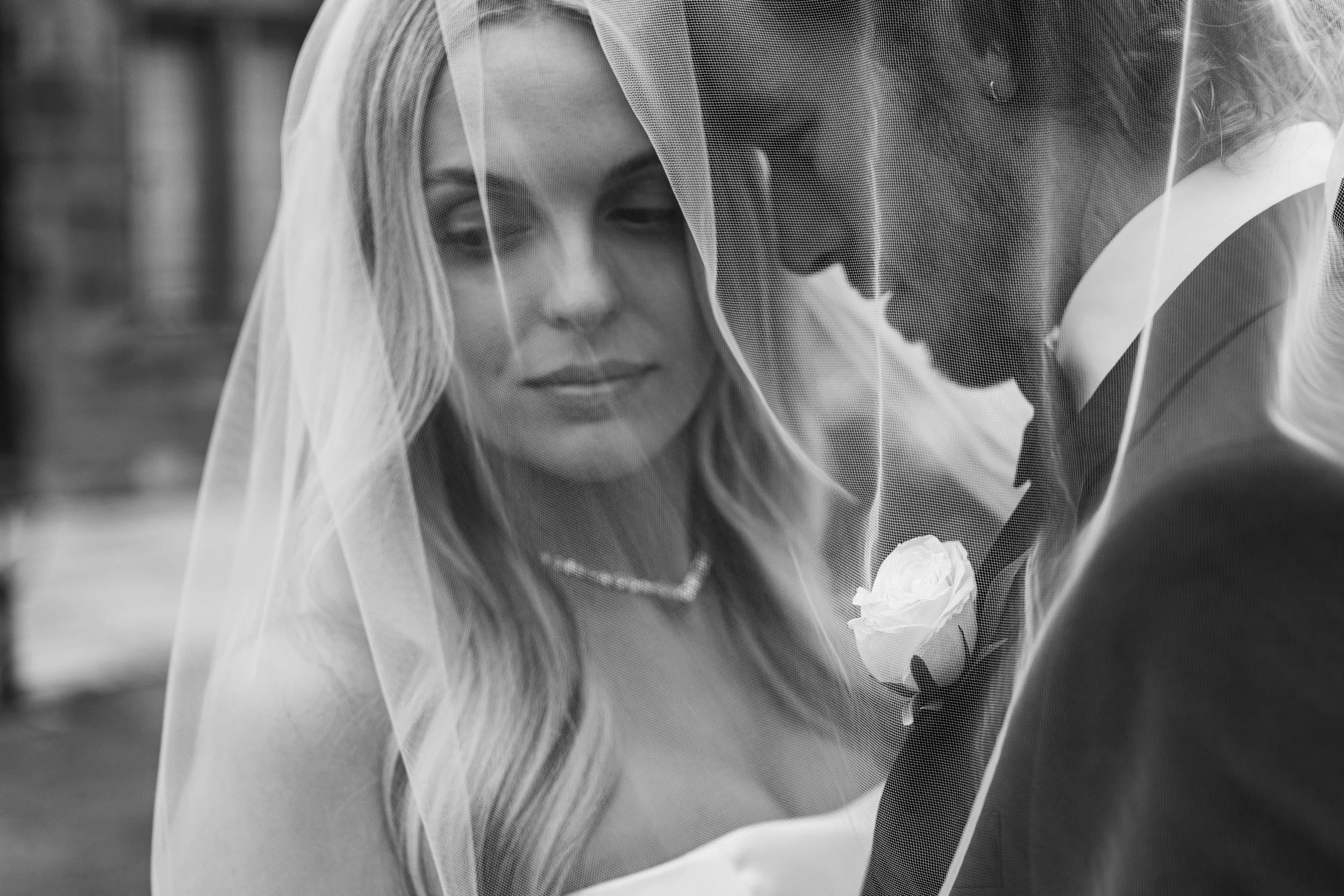 A black and white photo of a bride and groom close together, with the bride wearing a veil and pearl necklace, and the groom in a tuxedo with a white rose boutonniere, sharing an intimate moment.