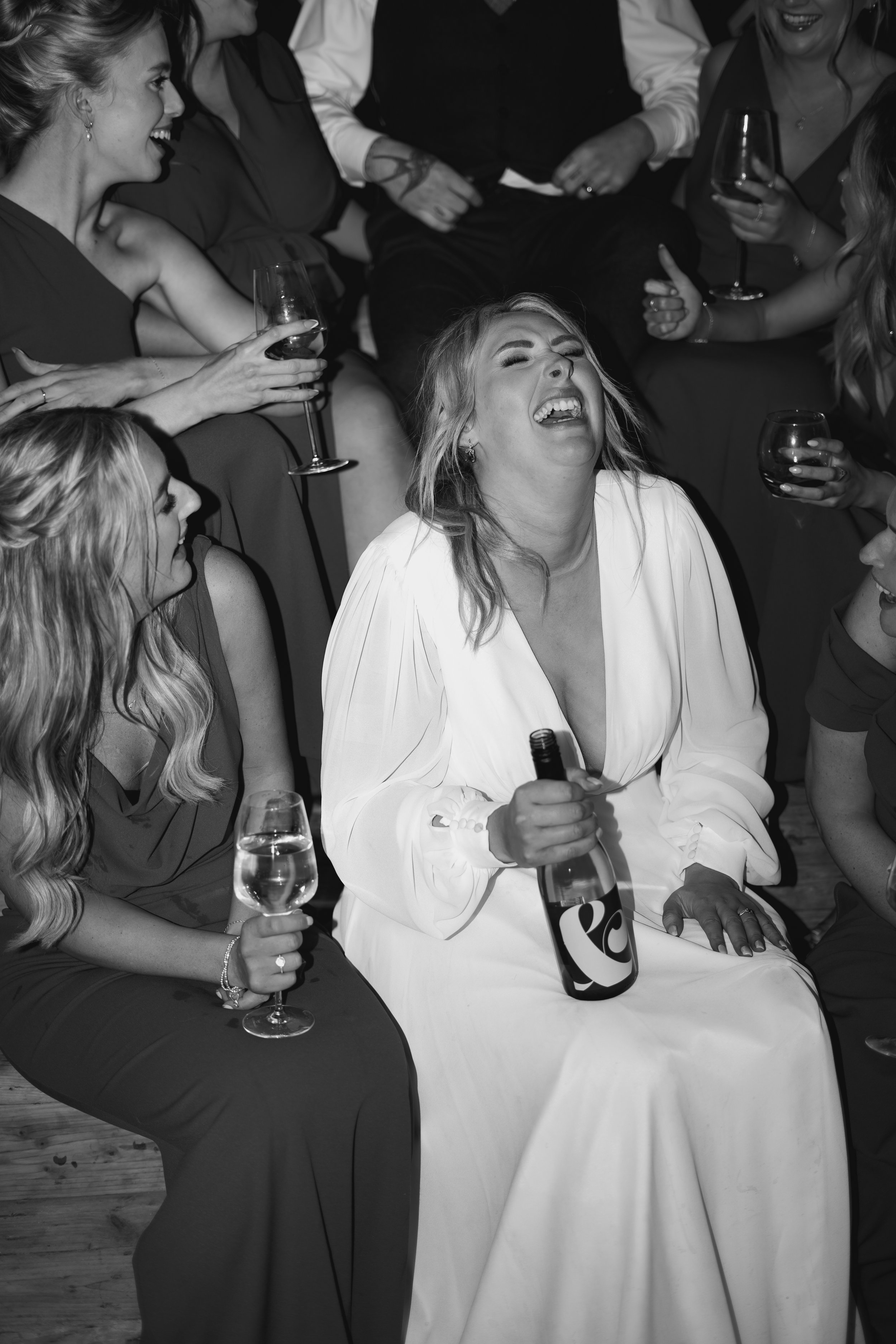 Group of women at a wedding, laughing and holding glasses of wine, with one woman in a white dress holding a bottle of wine, all sharing a joyful moment.