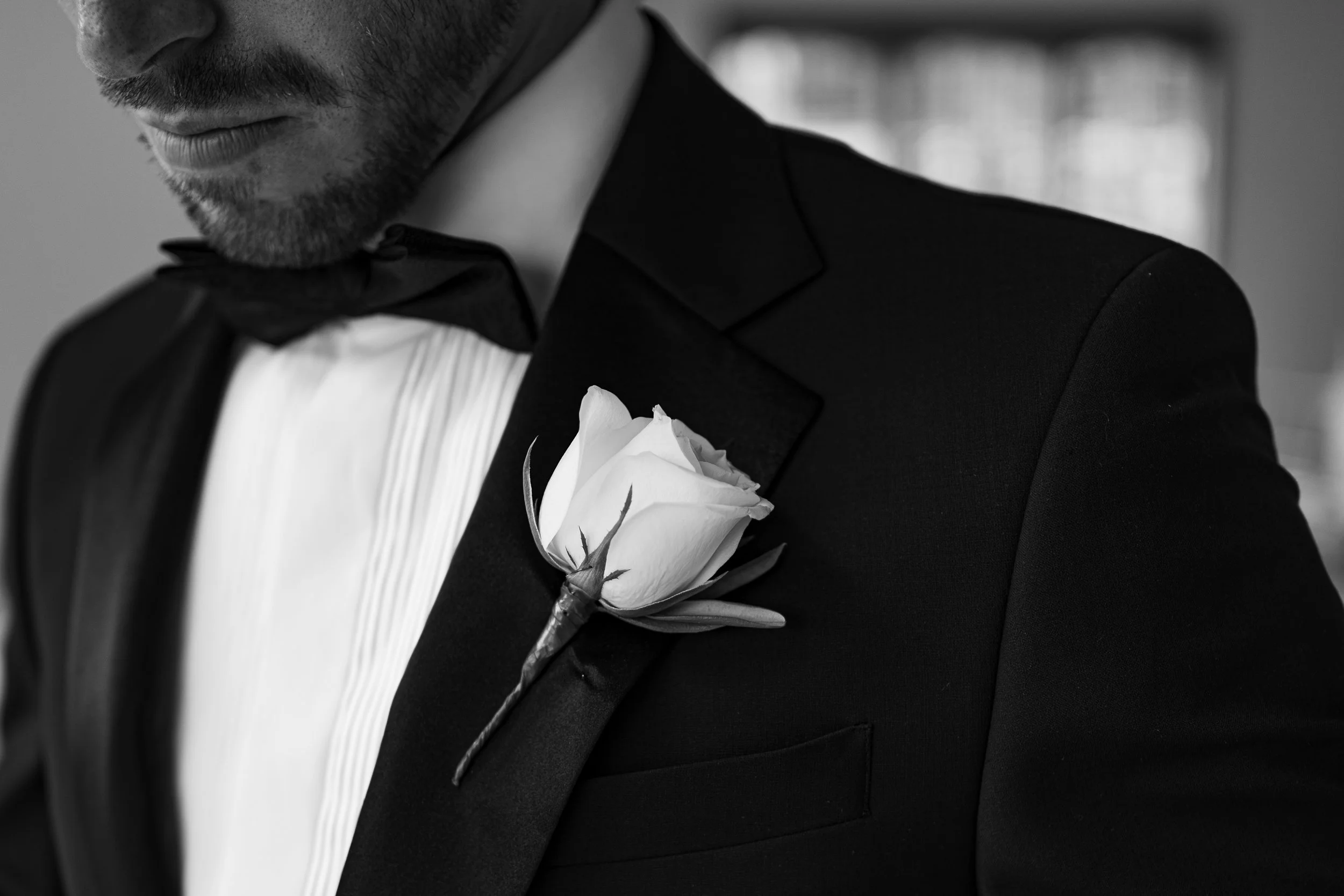 A man in formal attire, wearing a tuxedo with a white dress shirt and black bow tie, has a boutonniere made of a white rose pinned to his lapel.