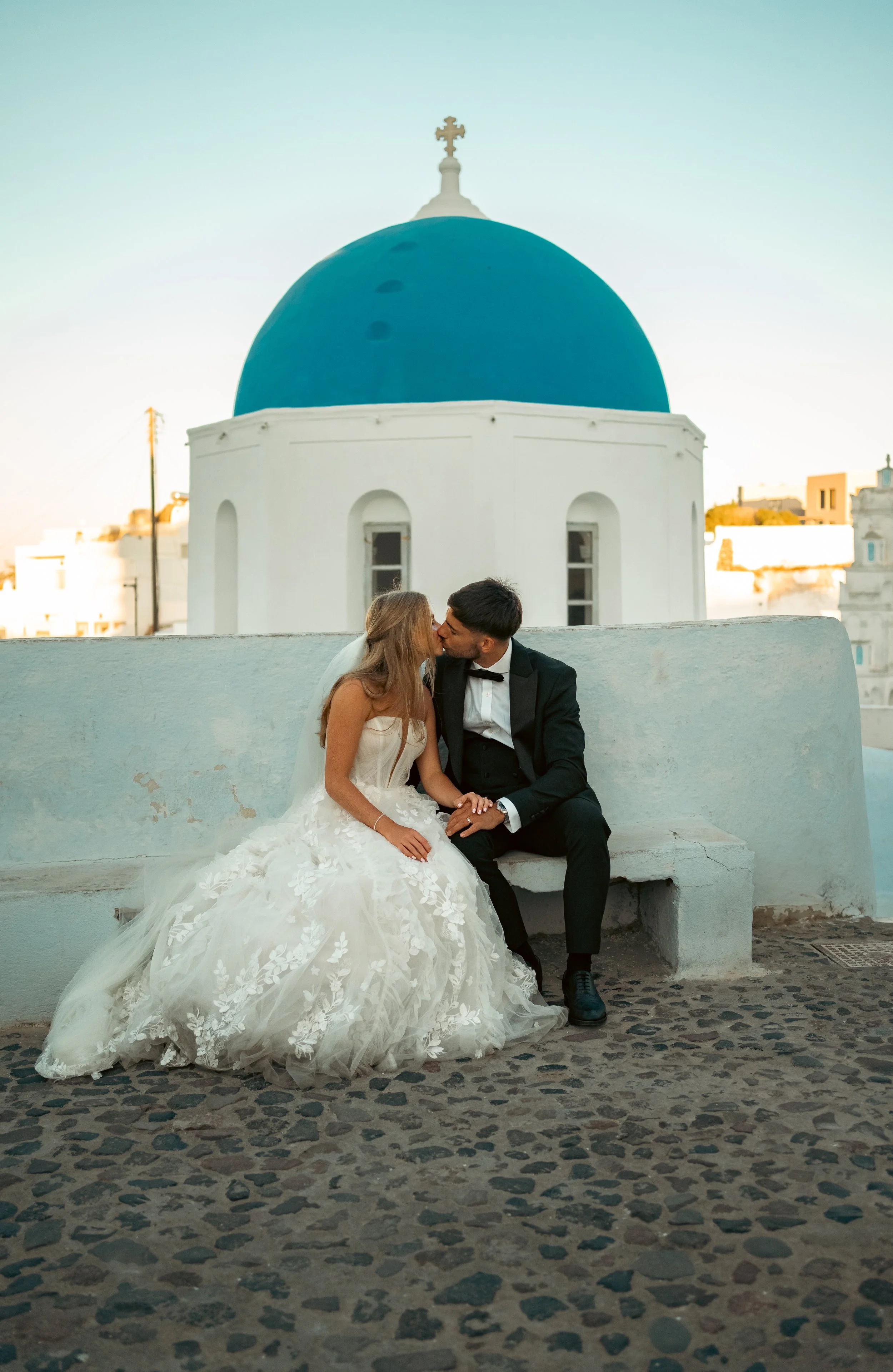 A bride and groom sharing a kiss on a white stone bench in front of a white building with a blue dome, likely in Greece.
