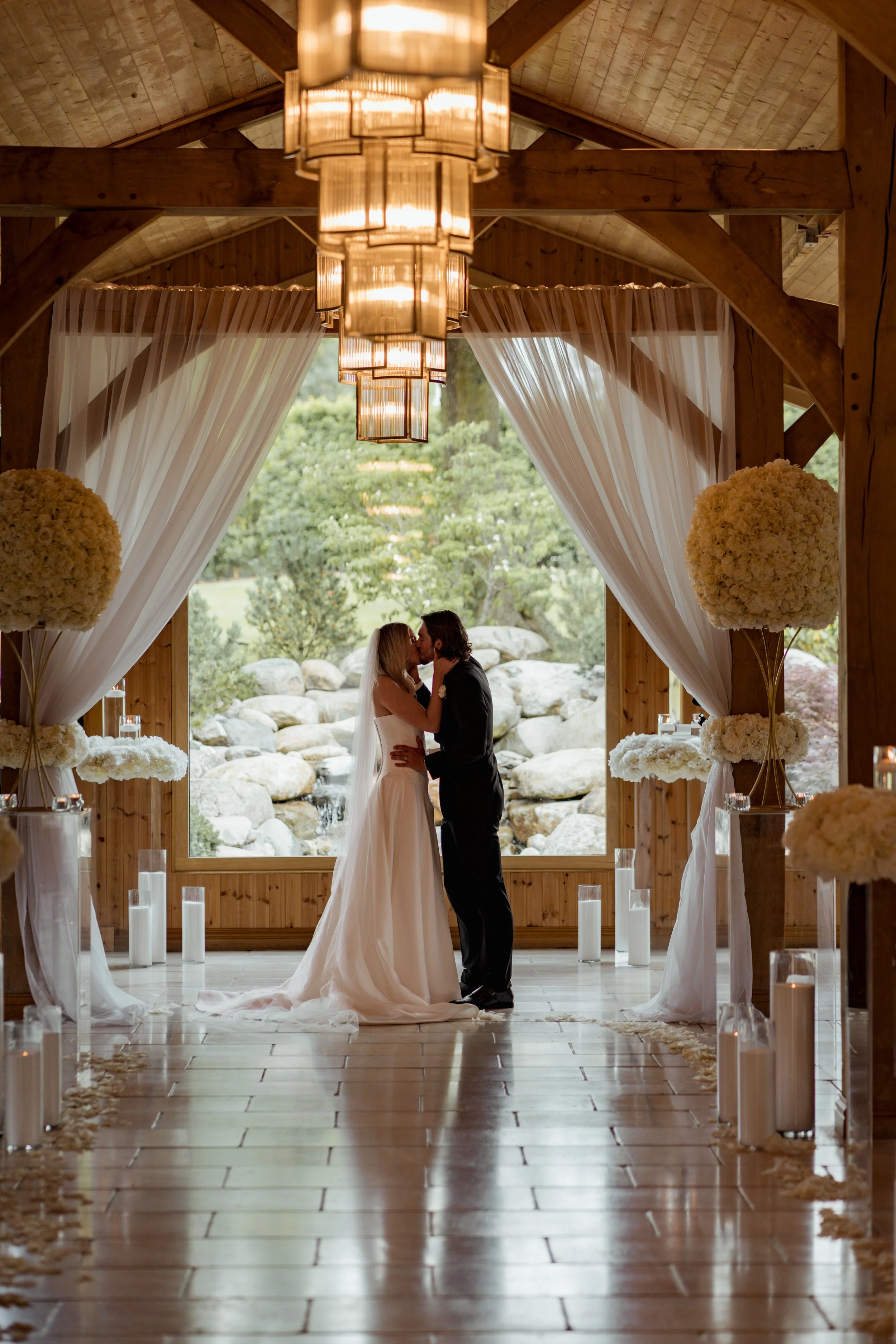 A bride and groom kissing at the altar of a decorated wedding venue with large floral arrangements and a backdrop of outdoors with trees and rocks.