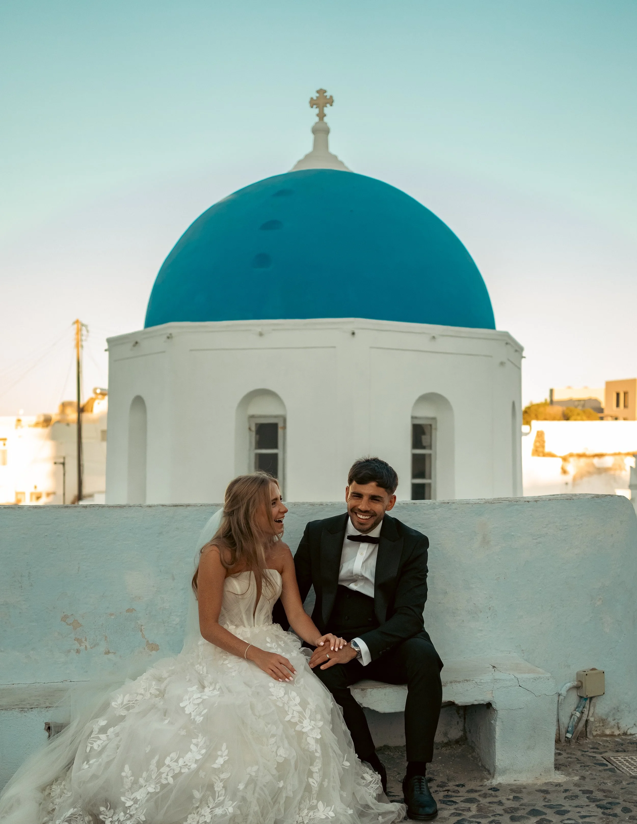 A happy bride and groom sitting on a white bench outside a building with a blue dome and white walls, likely in Greece. The bride is in a wedding dress, and the groom is in a tuxedo, both smiling and holding hands.