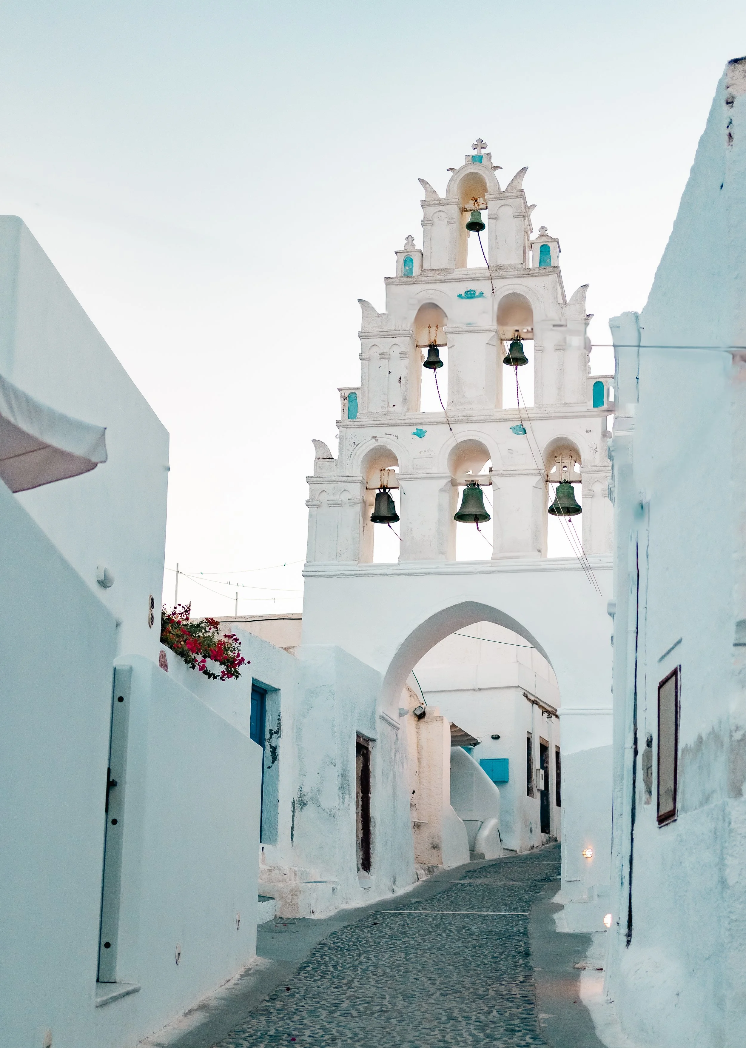 White church tower with multiple bells and crosses, situated in a narrow cobblestone street with whitewashed buildings, in Santorini.