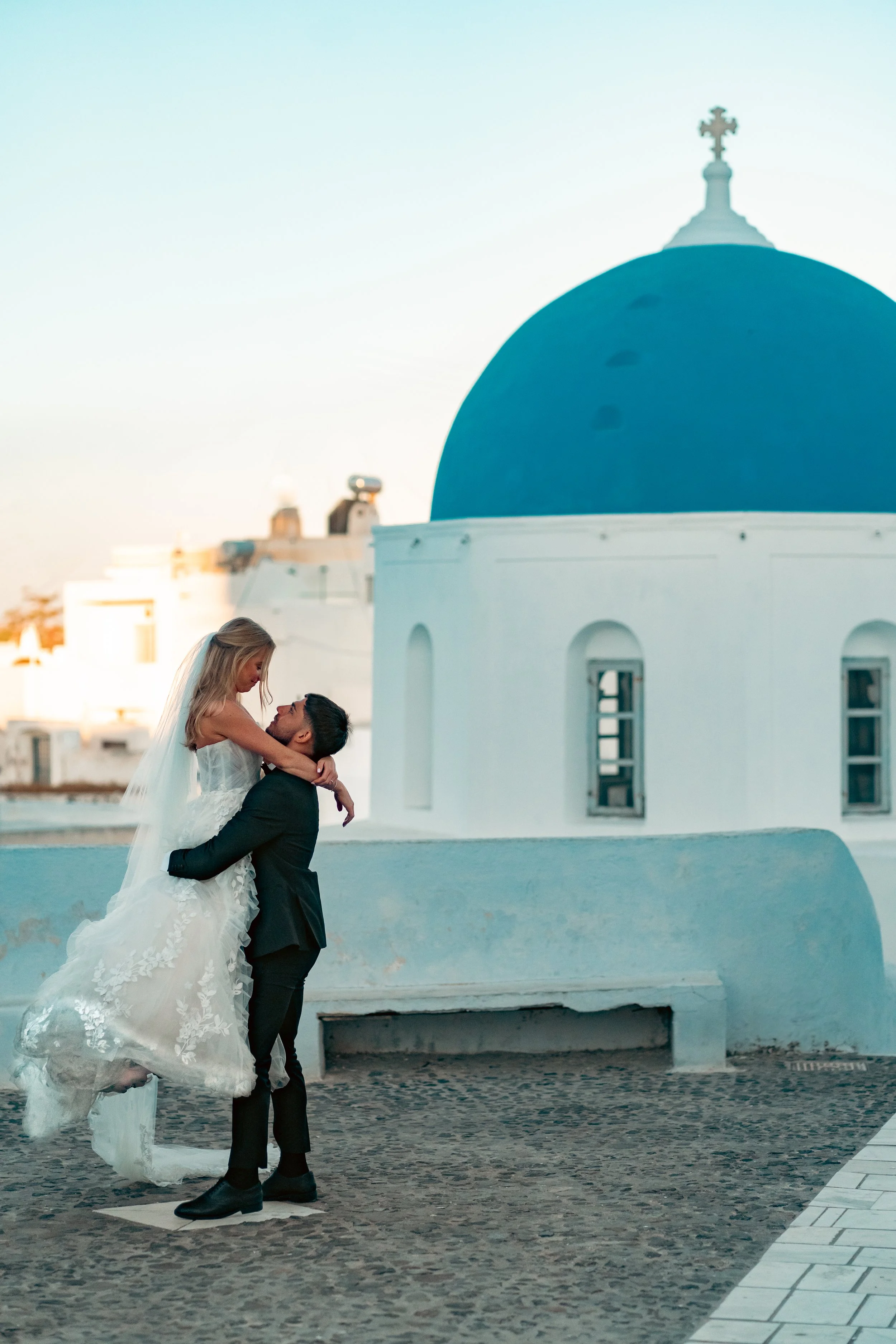 A bride being lifted by a groom on a cobblestone street in front of a white building with a blue-domed roof during sunrise in Santorini.