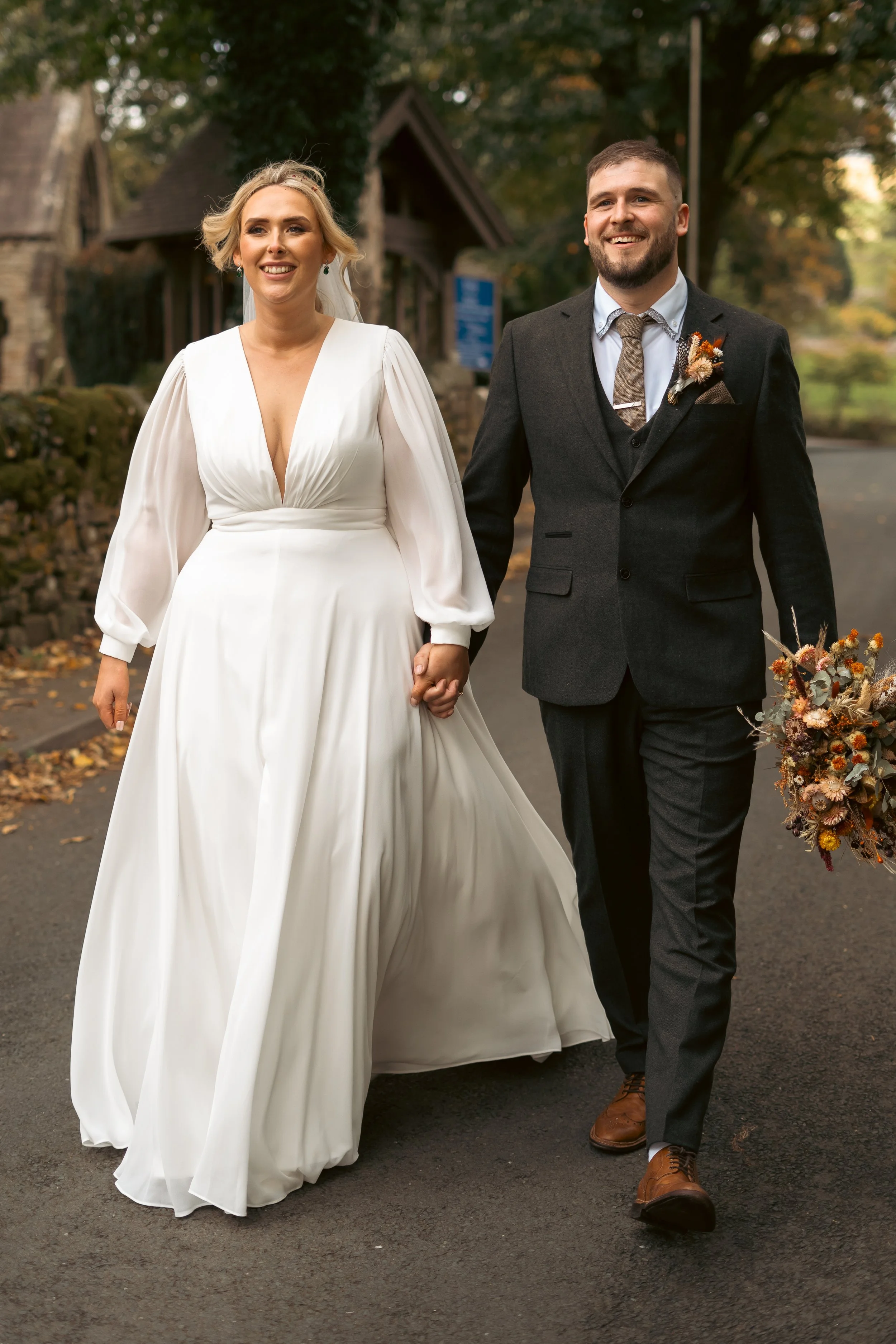 A newlywed couple holding hands and walking outdoors on a fall day, smiling. The bride is wearing a long white wedding dress with puffed sleeves, and the groom is wearing a dark suit with a tie, pocket square, and boutonniere. The background includes