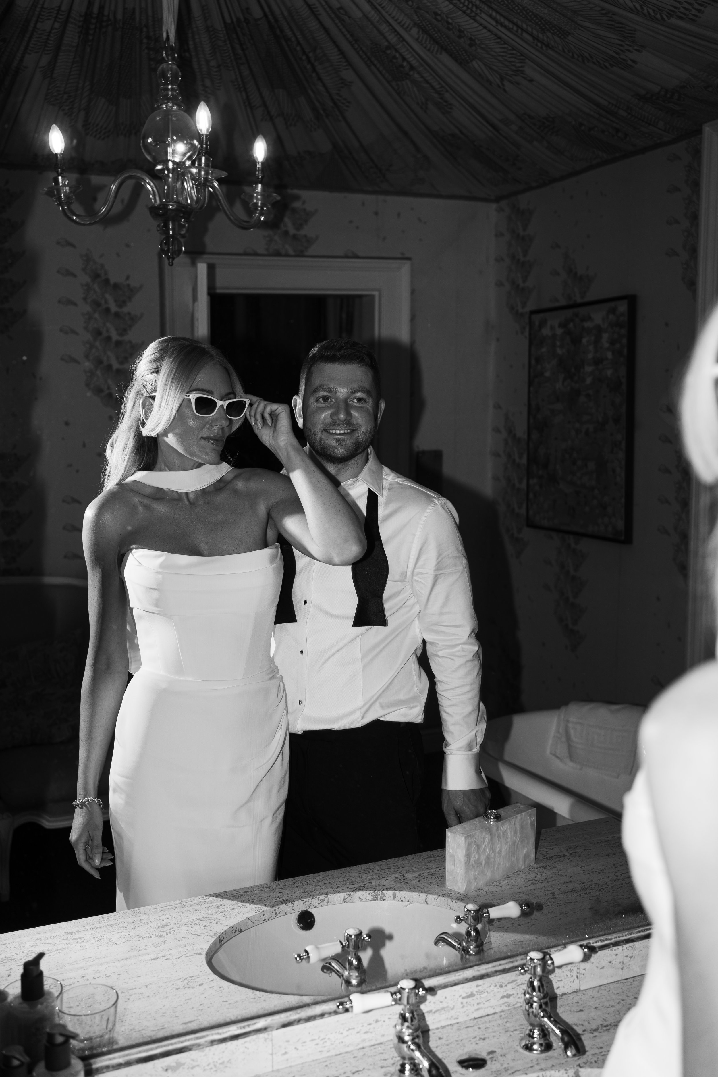 A woman in a white strapless dress and sunglasses, and a man in a dress shirt with a black bow tie hanging around his neck, standing in front of a mirror in a bathroom. The woman appears to be adjusting her sunglasses while the man is smiling. The room has a chandelier and framed wallpaper.