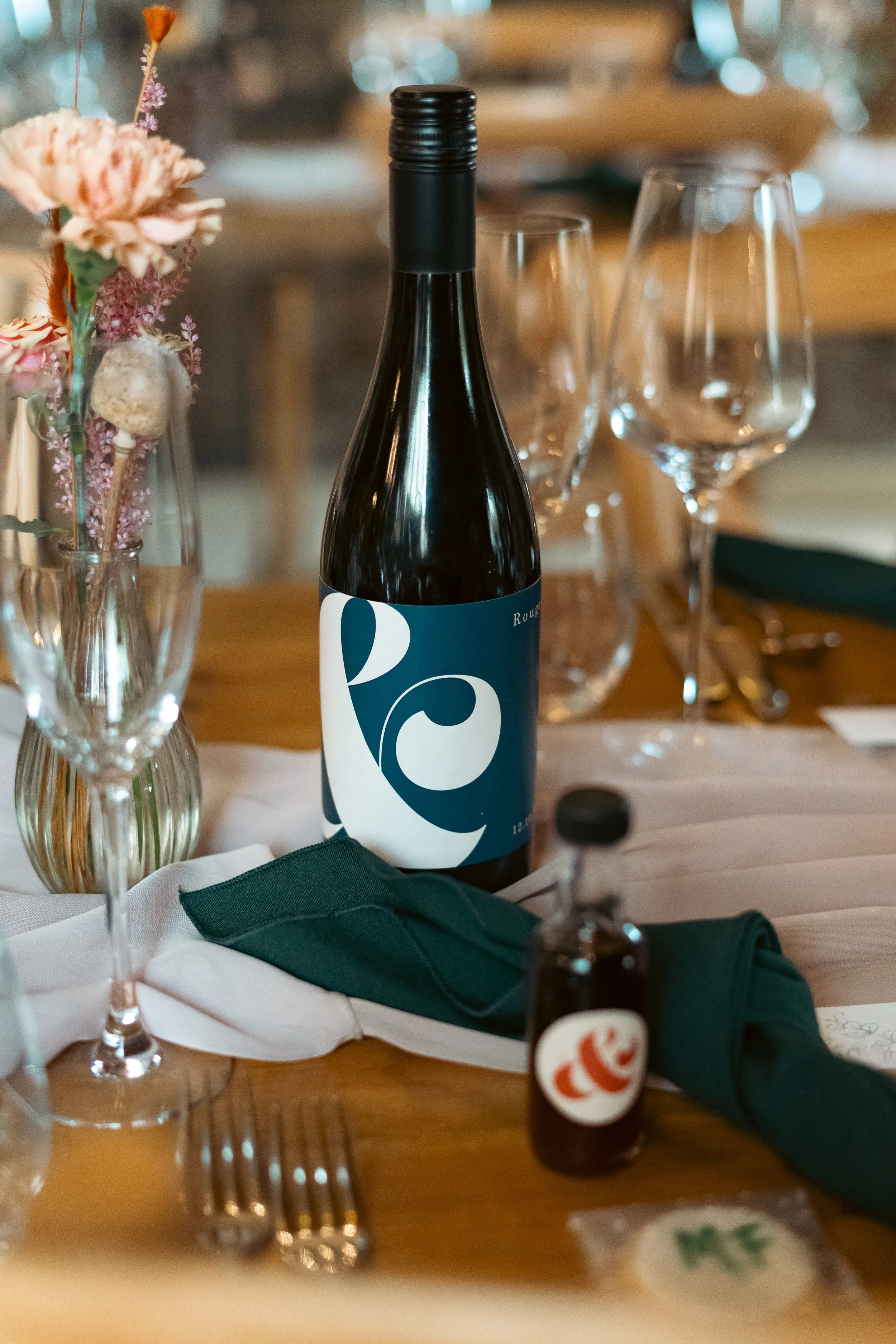 A table setting with a bottle of wine, wine glasses, a small vase with dried flowers, a green cloth napkin, and a small bottle of hot sauce at a rustic dining event.