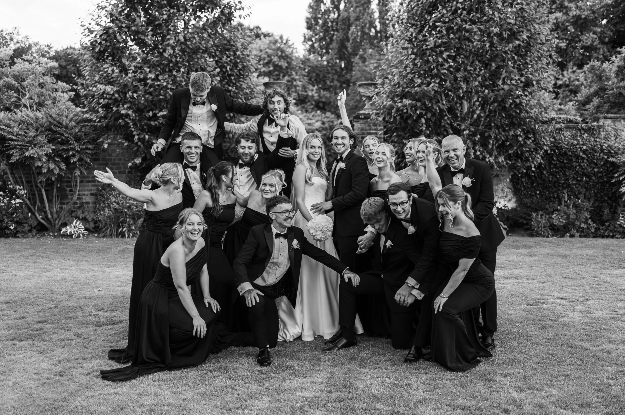 A wedding party in formal attire posing and making funny gestures outdoors in a garden.