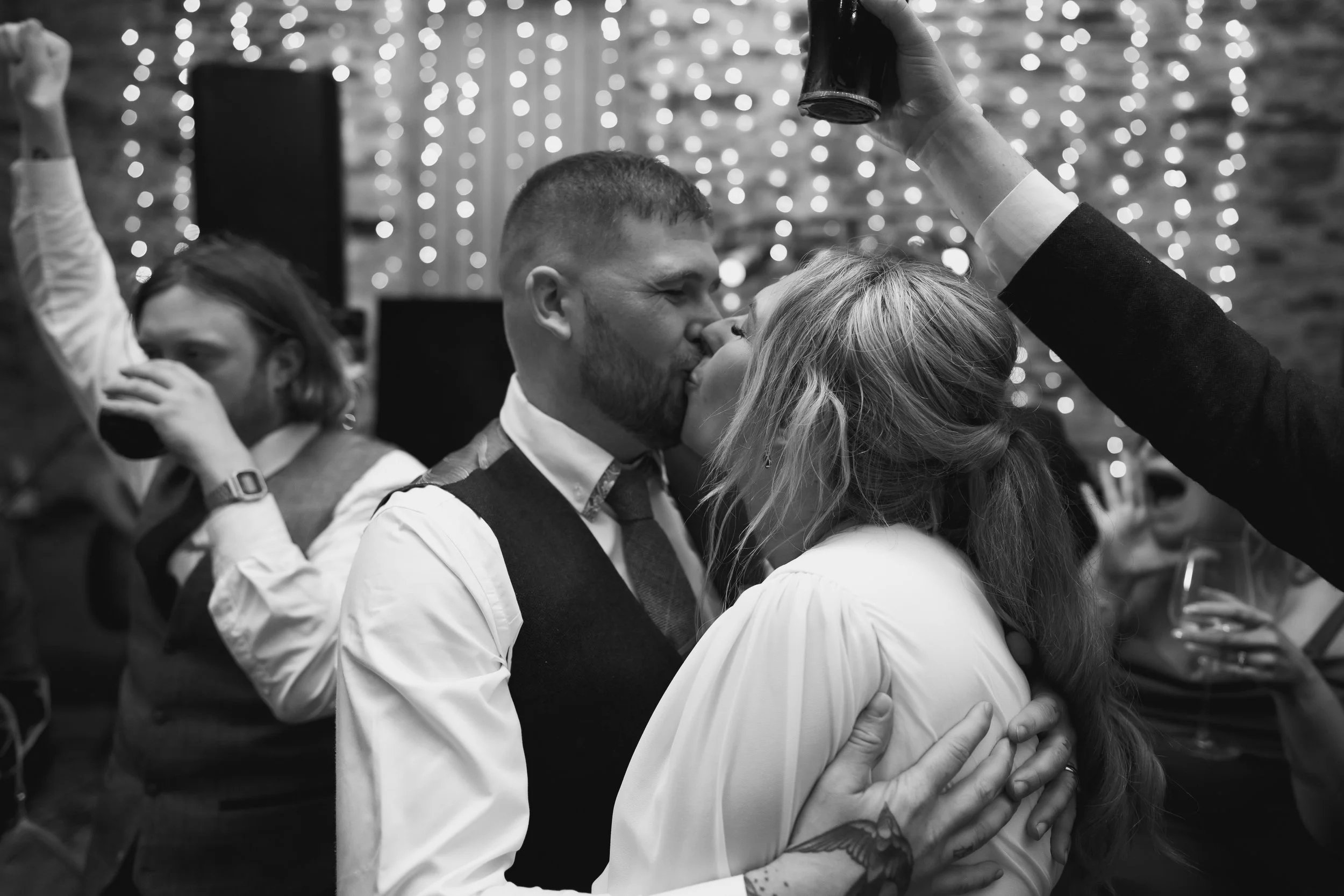 A couple kisses at a celebration, with a man in a vest and a woman with loose hair embracing and kissing. In the background, a man drinks from a glass, and a woman holds a drink. Decorative string lights hang behind them.