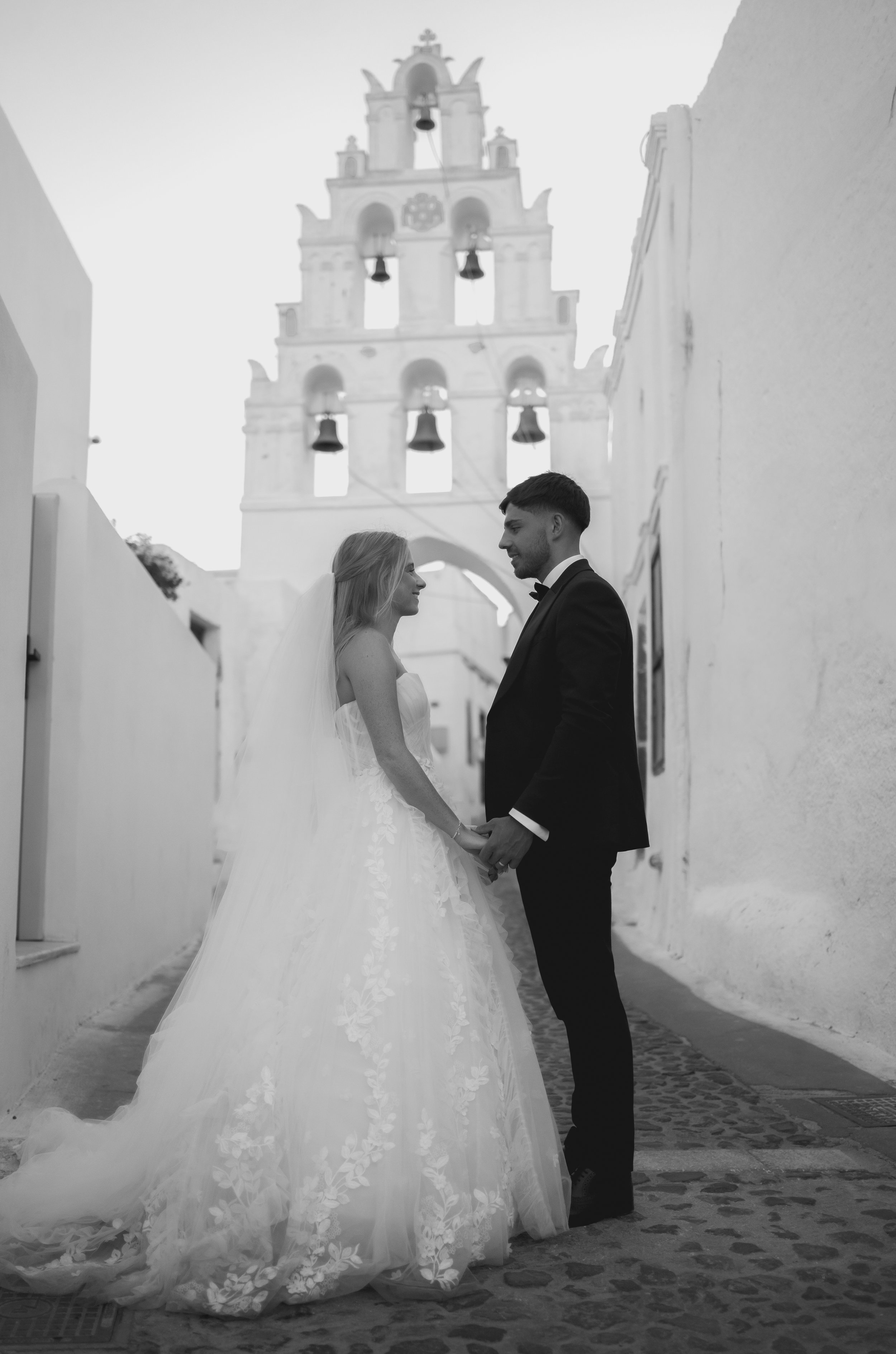 Black and white photo of a bride and groom holding hands and facing each other in a narrow street with a church bell tower in the background, during their wedding in Santorini.