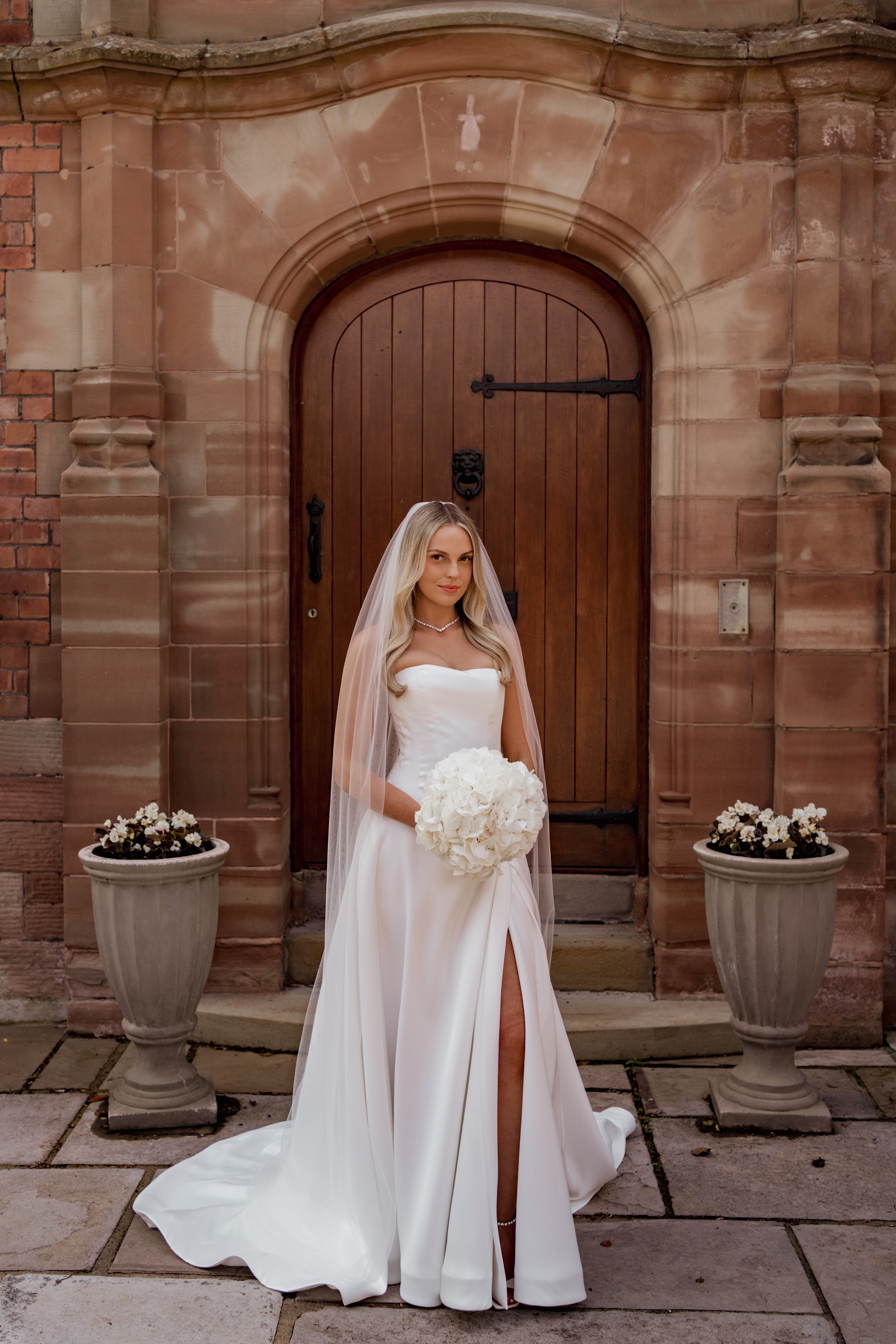 A bride in a white wedding gown and veil holding a bouquet of white flowers standing in front of a wooden door with stone accents, flanked by two flower pots with white flowers on either side.