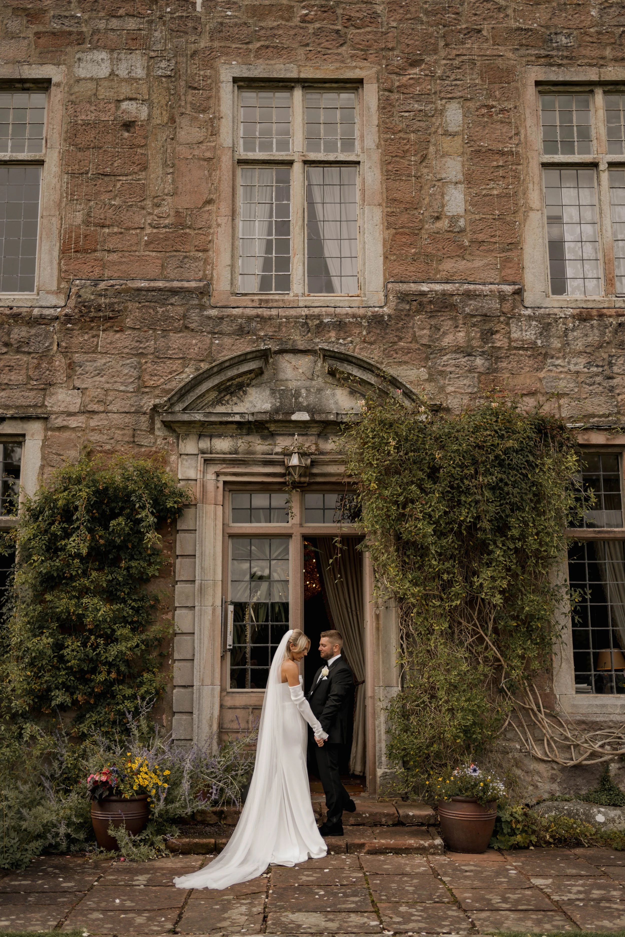 Bride and groom holding hands outside an old stone building with large windows and greenery, on a brick patio.