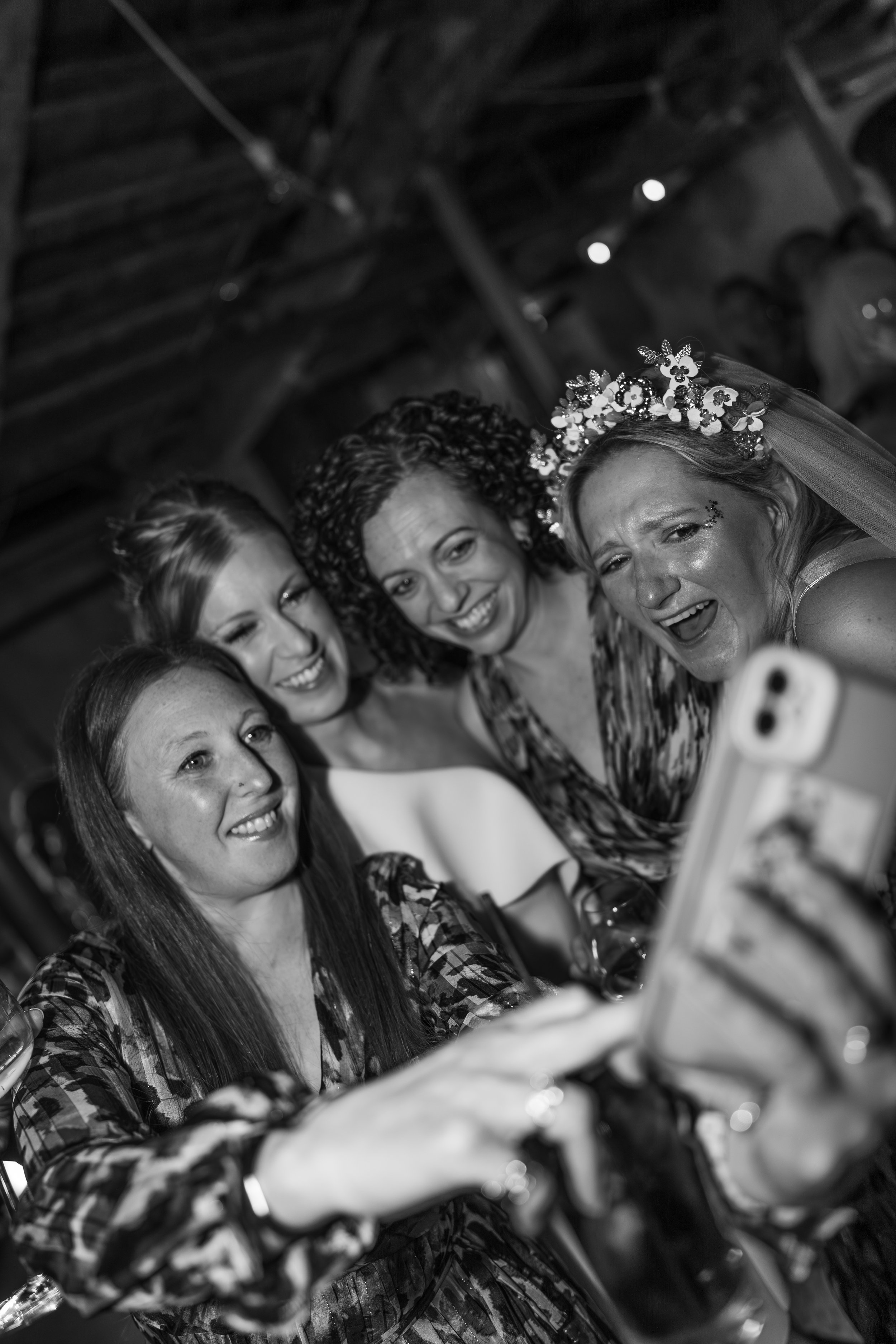 Five women taking a selfie at a celebration, with one woman wearing a floral headpiece and veil, smiling and looking at a smartphone camera.