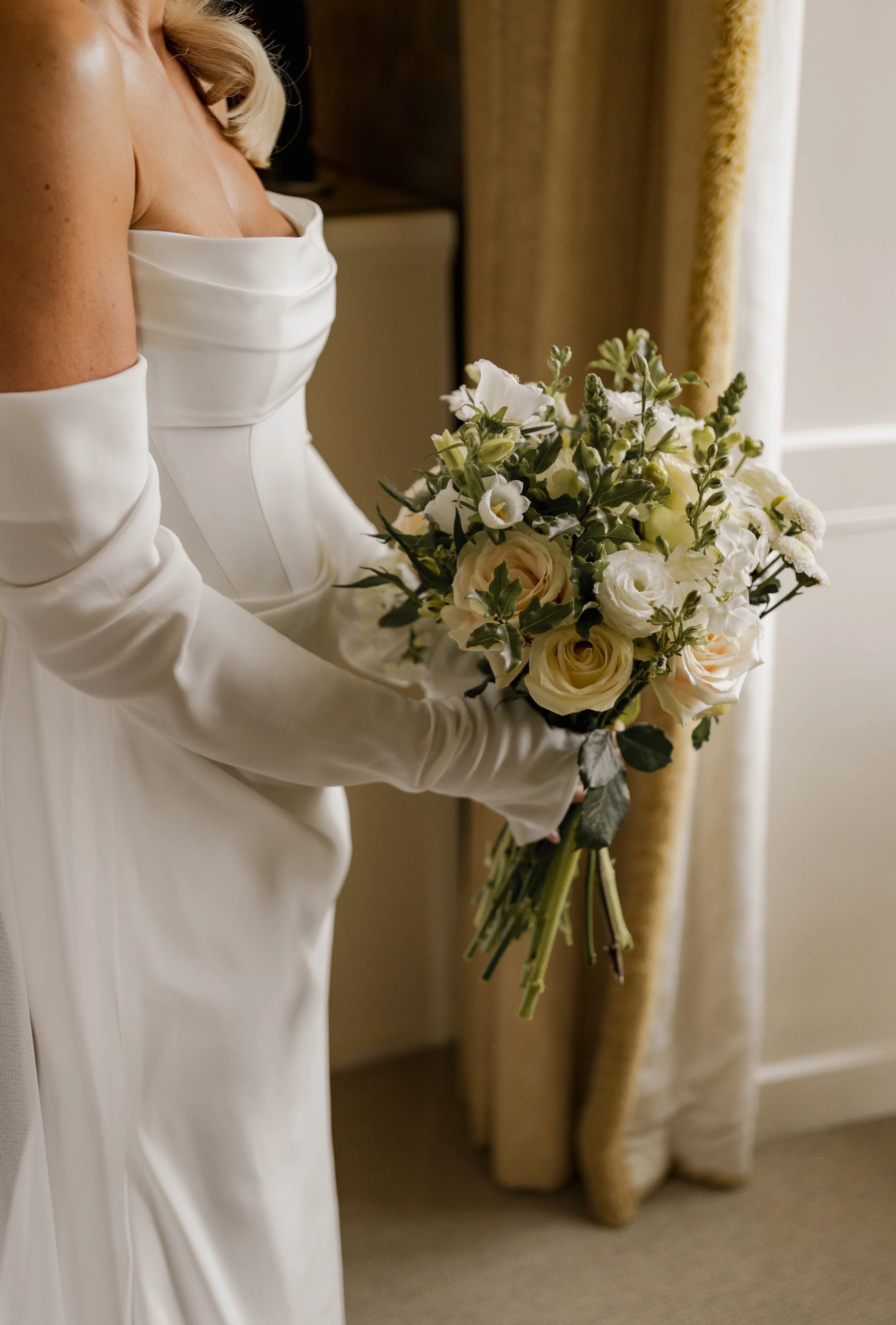 A bride in a white satin wedding dress and long gloves holding a bouquet of white and cream roses and other flowers indoors at Askham Hall