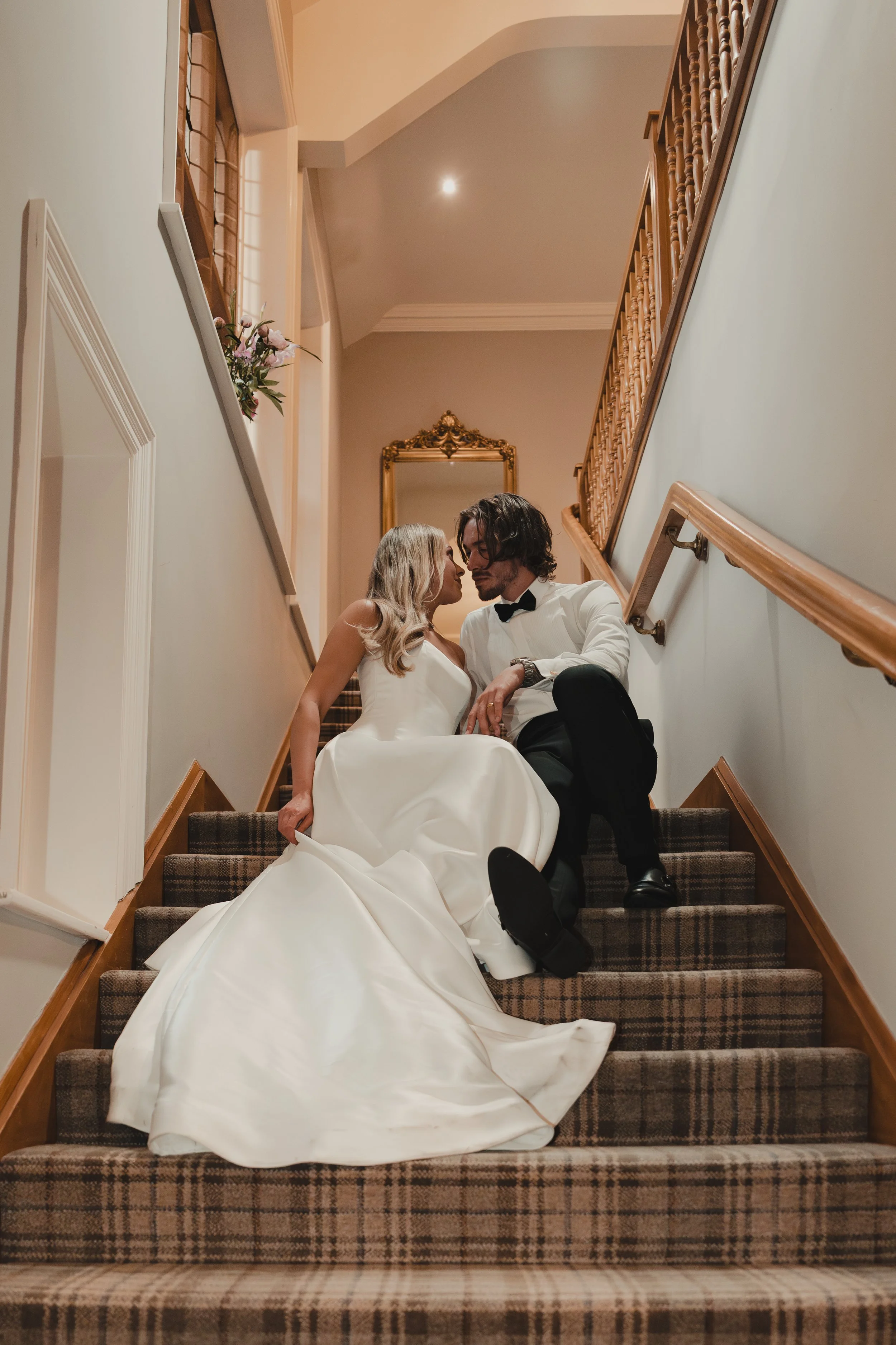 A bride and groom sitting on a staircase inside a building, dressed in wedding attire, gazing at each other intimately.