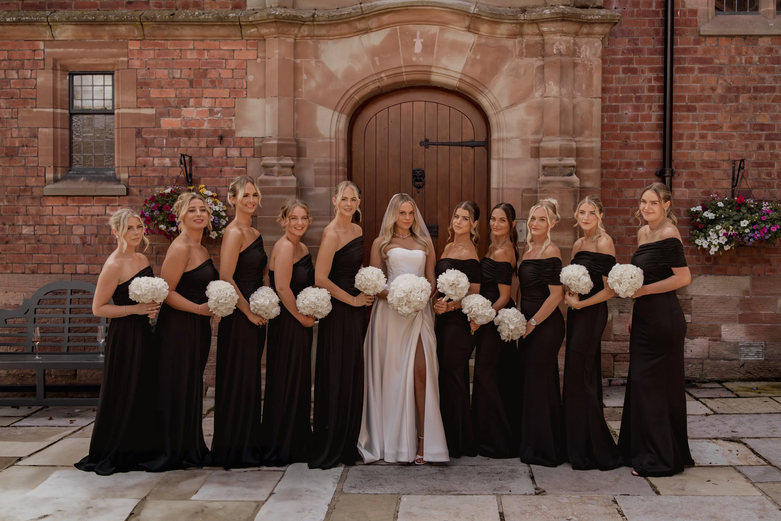 A group of eleven women, including a bride in a white wedding dress and ten bridesmaids in black dresses, standing in front of a brick building with floral arrangements.