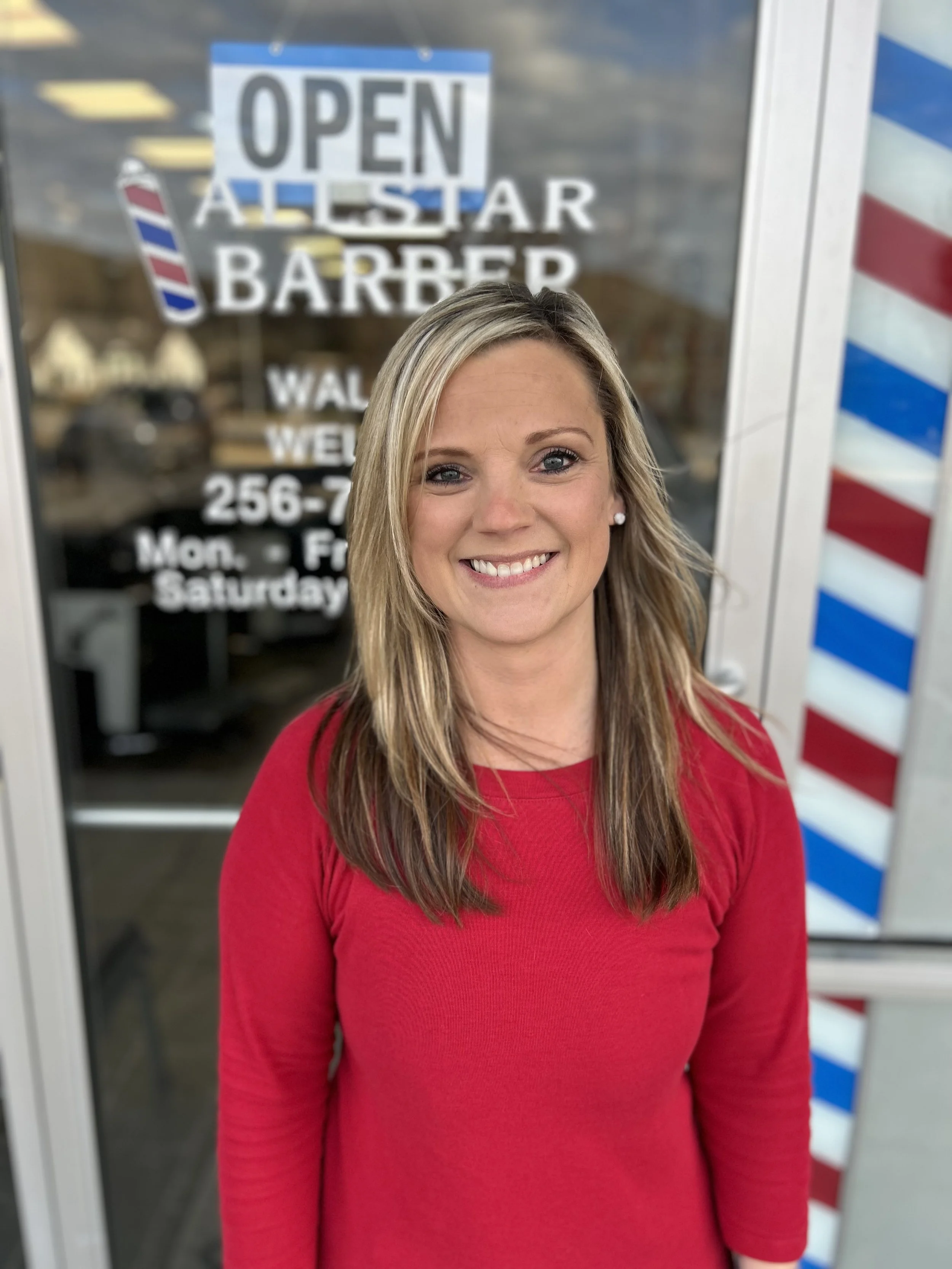 Woman with blonde hair wearing a red shirt, smiling outdoors in front of a barbershop window with a sign that says 'Open All Star Barber'.