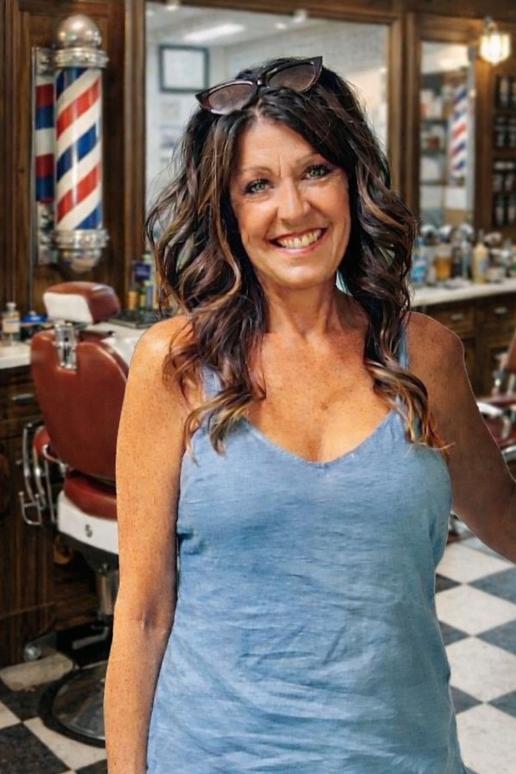 Smiling woman with wavy brown hair, glasses on her head, wearing a blue sleeveless top in a vintage barbershop
