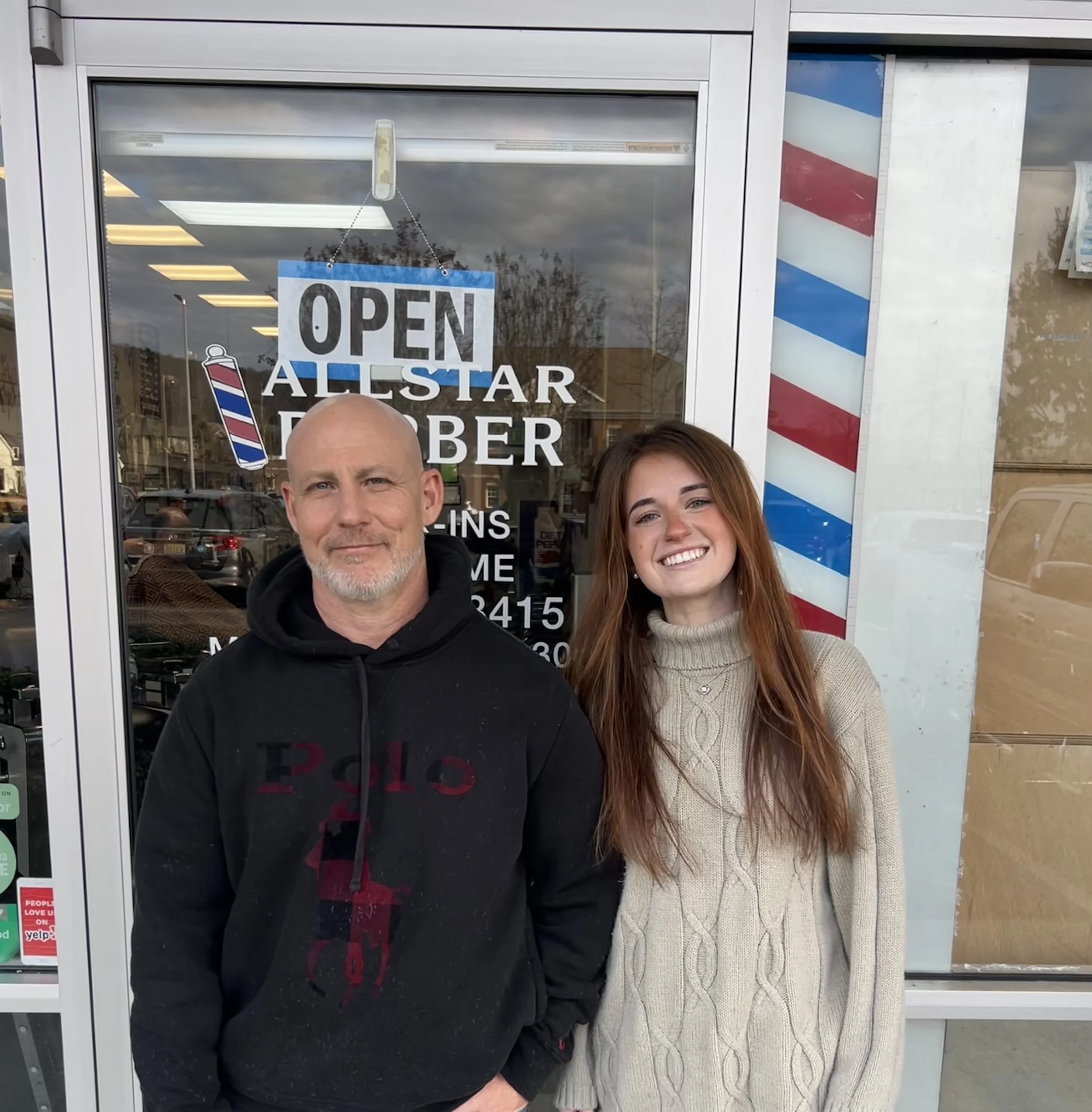 A man and a woman standing in front of a barber shop window with an 'Open' sign, barber pole decoration, and mirror inside.