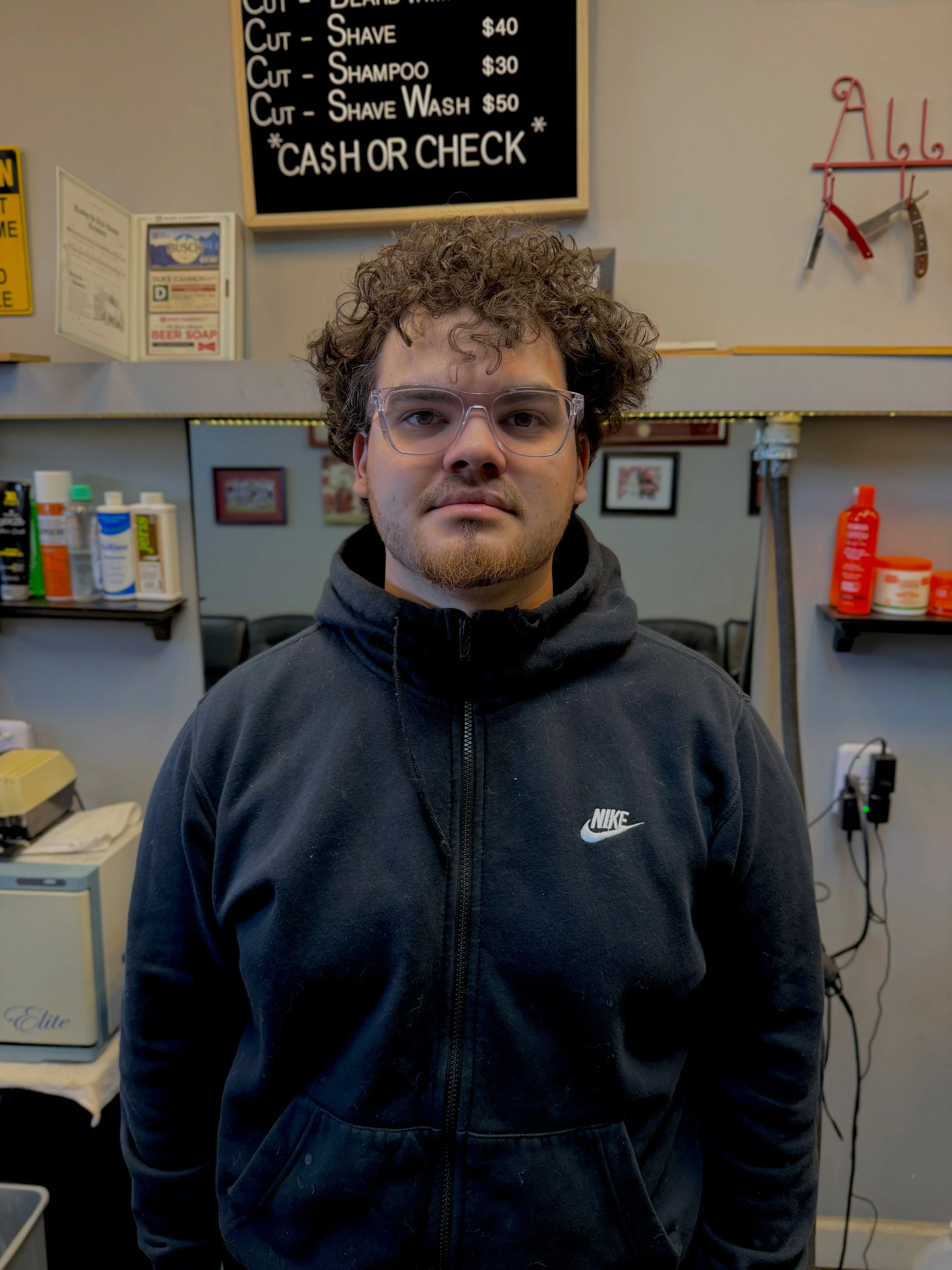 A young man with curly hair and glasses wearing a black Nike hoodie, standing in a barbershop or salon with grooming products on shelves in the background.