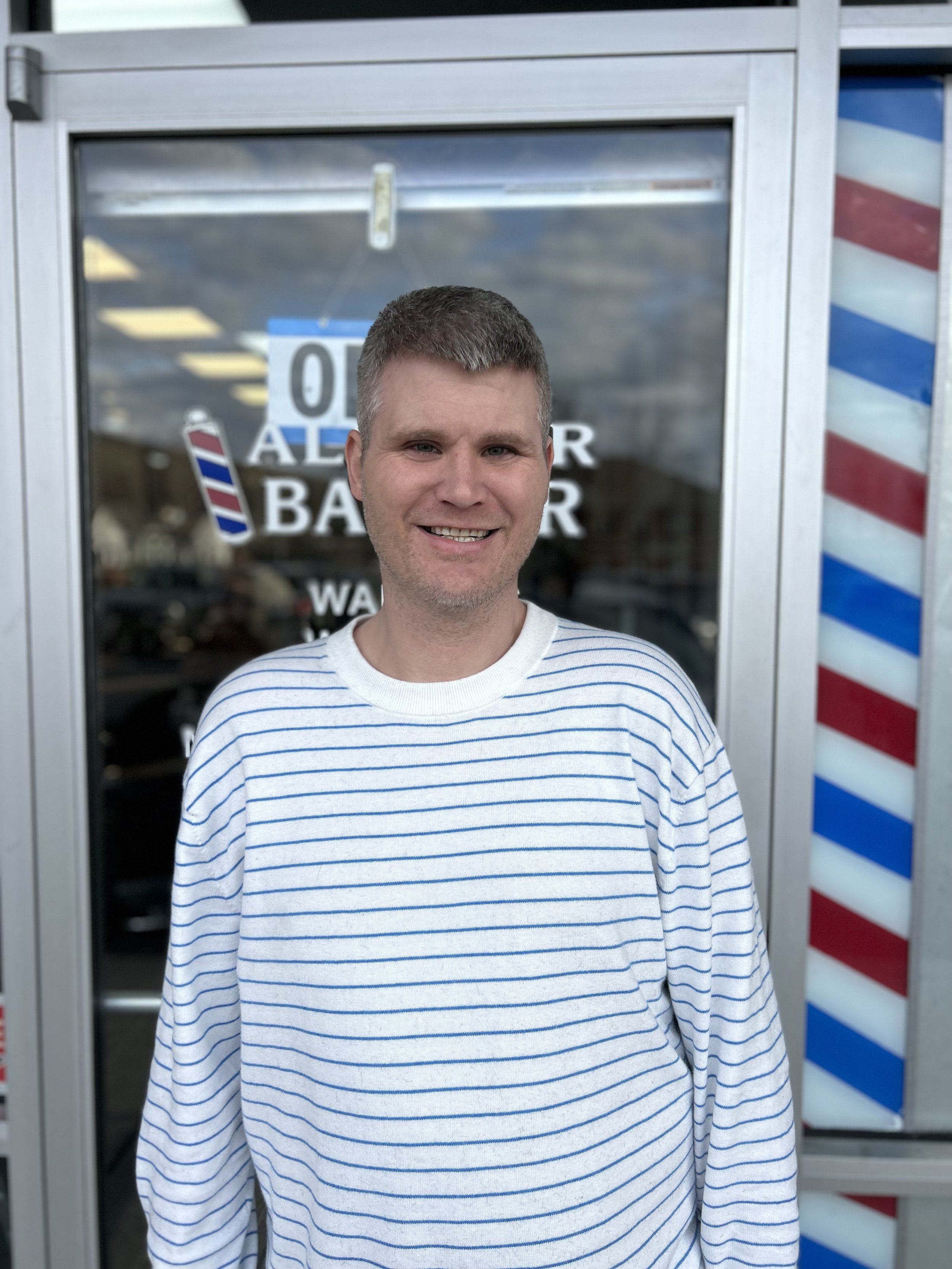 A man wearing a white and blue striped sweater standing outside a barber shop with a traditional barber pole in the background.