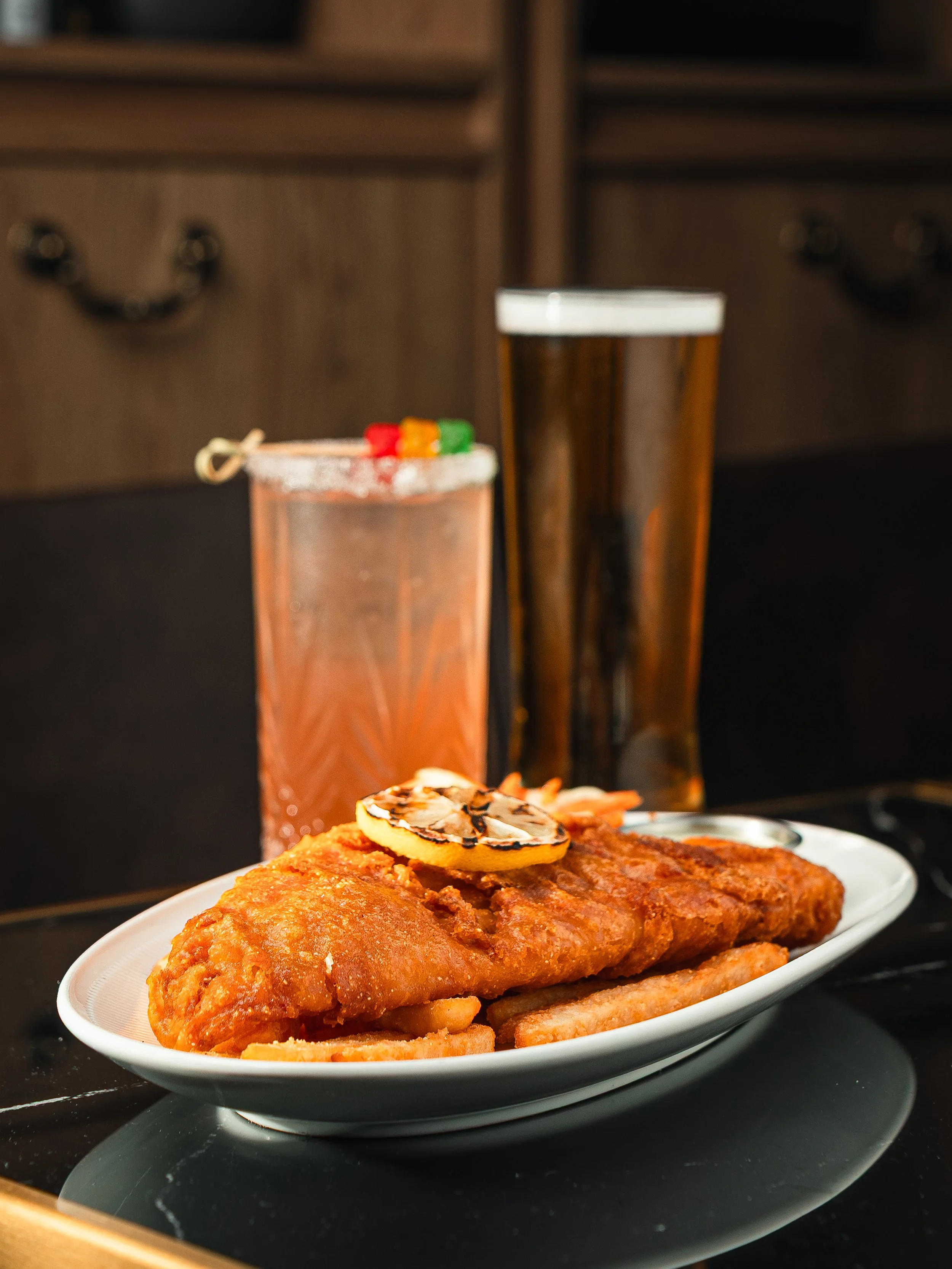 Fried fish fillet with a lemon slice on top, served on a white plate with French fries, alongside two glasses of dark-colored soft drinks.