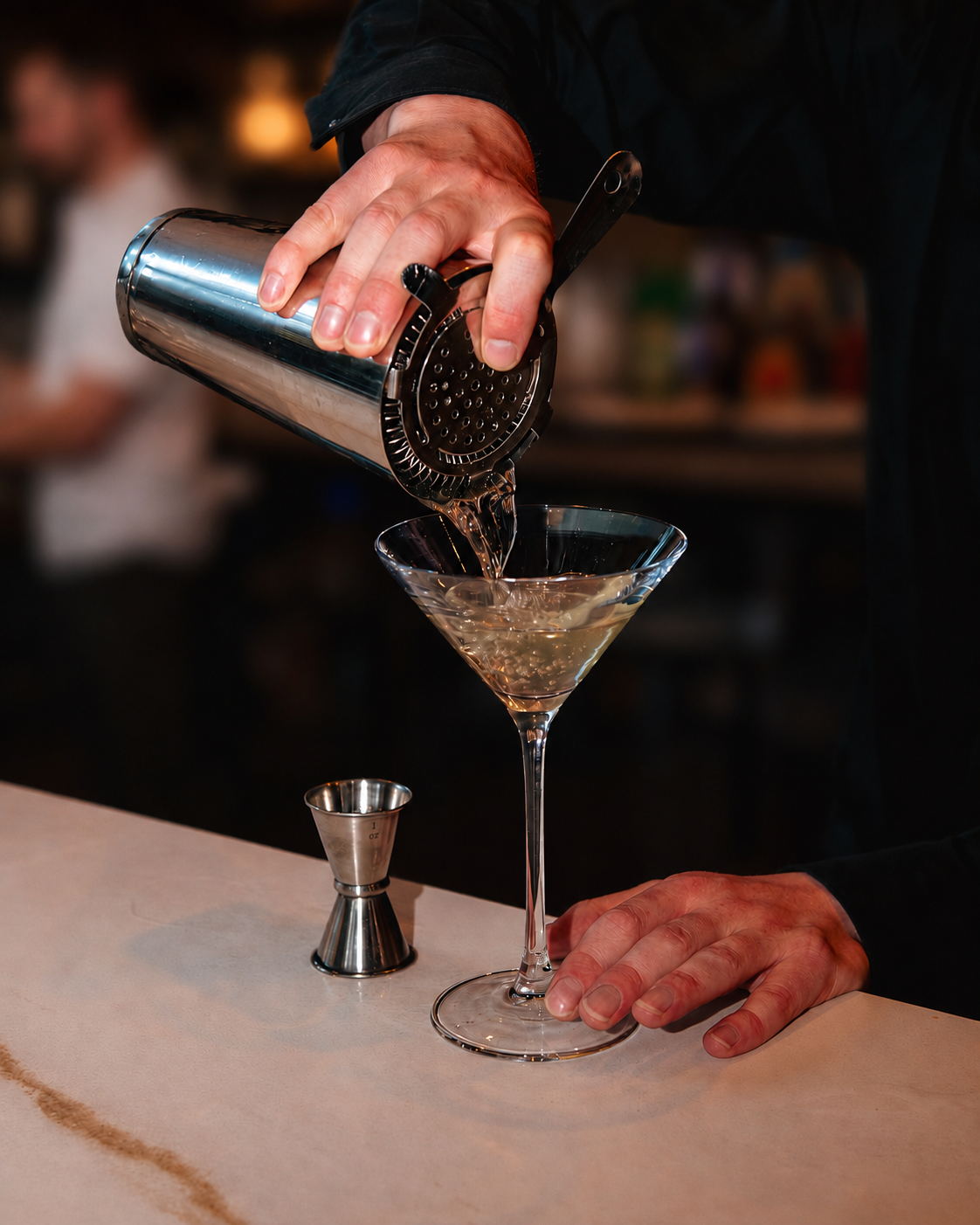 A bartender pouring a cocktail from a metal shaker into a martini glass, with a jigger on the bar counter.
