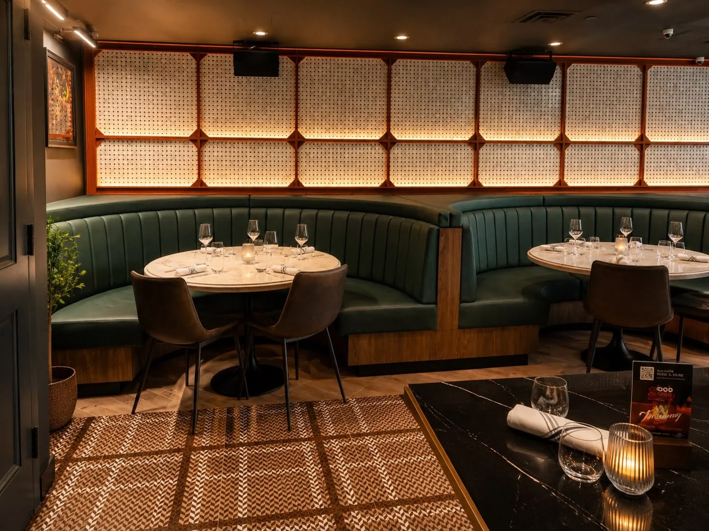 Elegant restaurant with curved green booth seating, round marble tables set with wine glasses, water glasses, and candles, and warm ambient lighting.