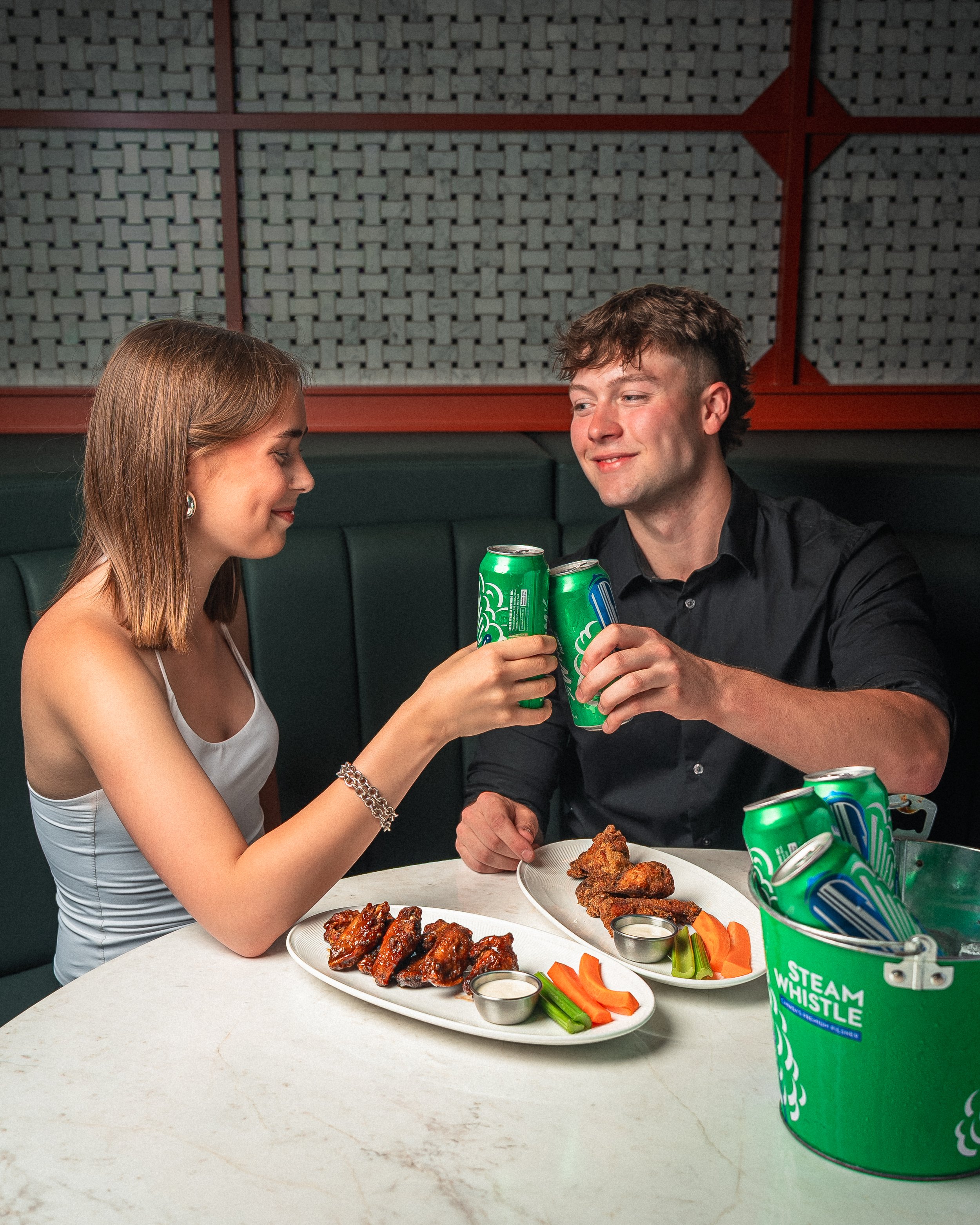 A young woman and a young man sitting at a table, clinking cans of Sprite soda. The table has plates of chicken wings, fried chicken, carrots, celery, and ranch dipping sauces. The setting appears to be a restaurant with a dark green booth and a decorative wall in the background.