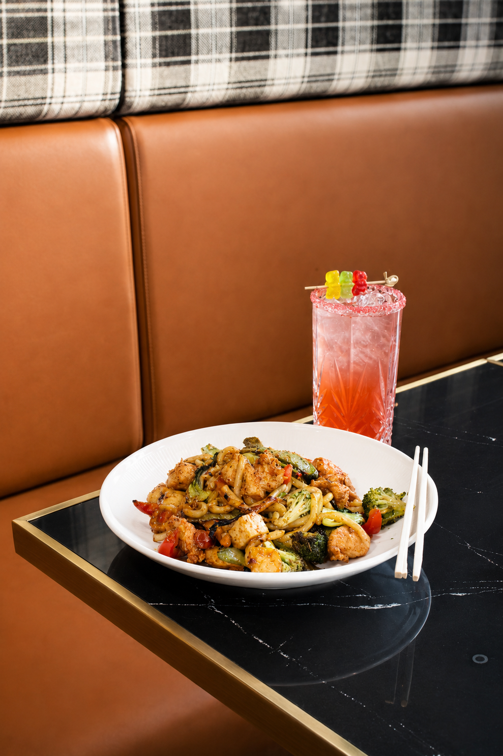 A plate of stir-fried noodles with vegetables and chicken on a black marble table with a pink cocktail garnished with gummy candies and a straw.