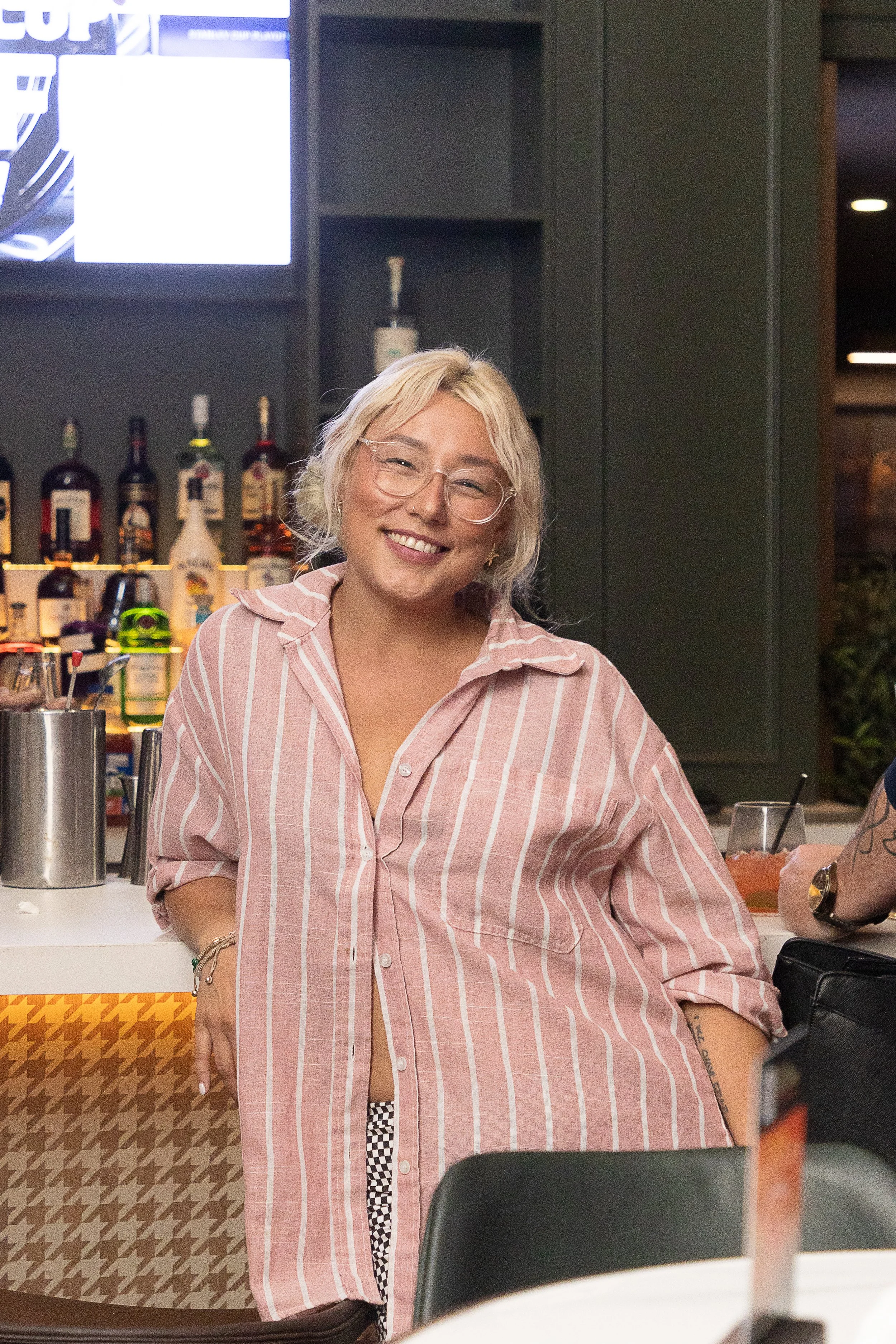 A woman with blonde hair, glasses, and a pink striped shirt smiling indoors. Behind her are bottles of alcohol on a shelf, a television screen, and a bar area.
