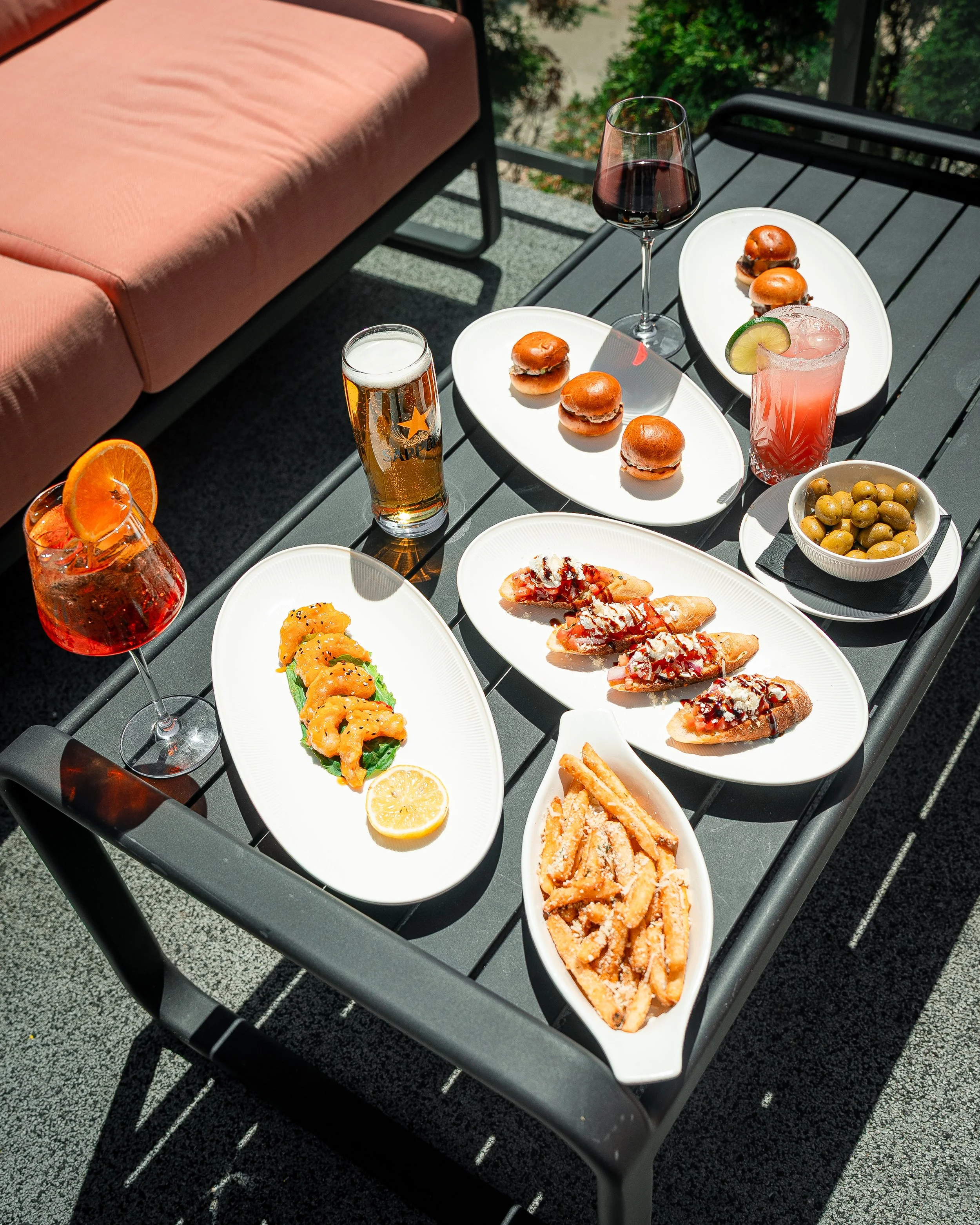 Outdoor patio table with various snacks and drinks. There are plates of sliders, a bowl of green olives, a glass of red wine, a glass of beer, a cocktail with a lime wedge, and a pizza slice. The table is on a deck with a pink cushion sofa nearby, and greenery can be seen in the background.