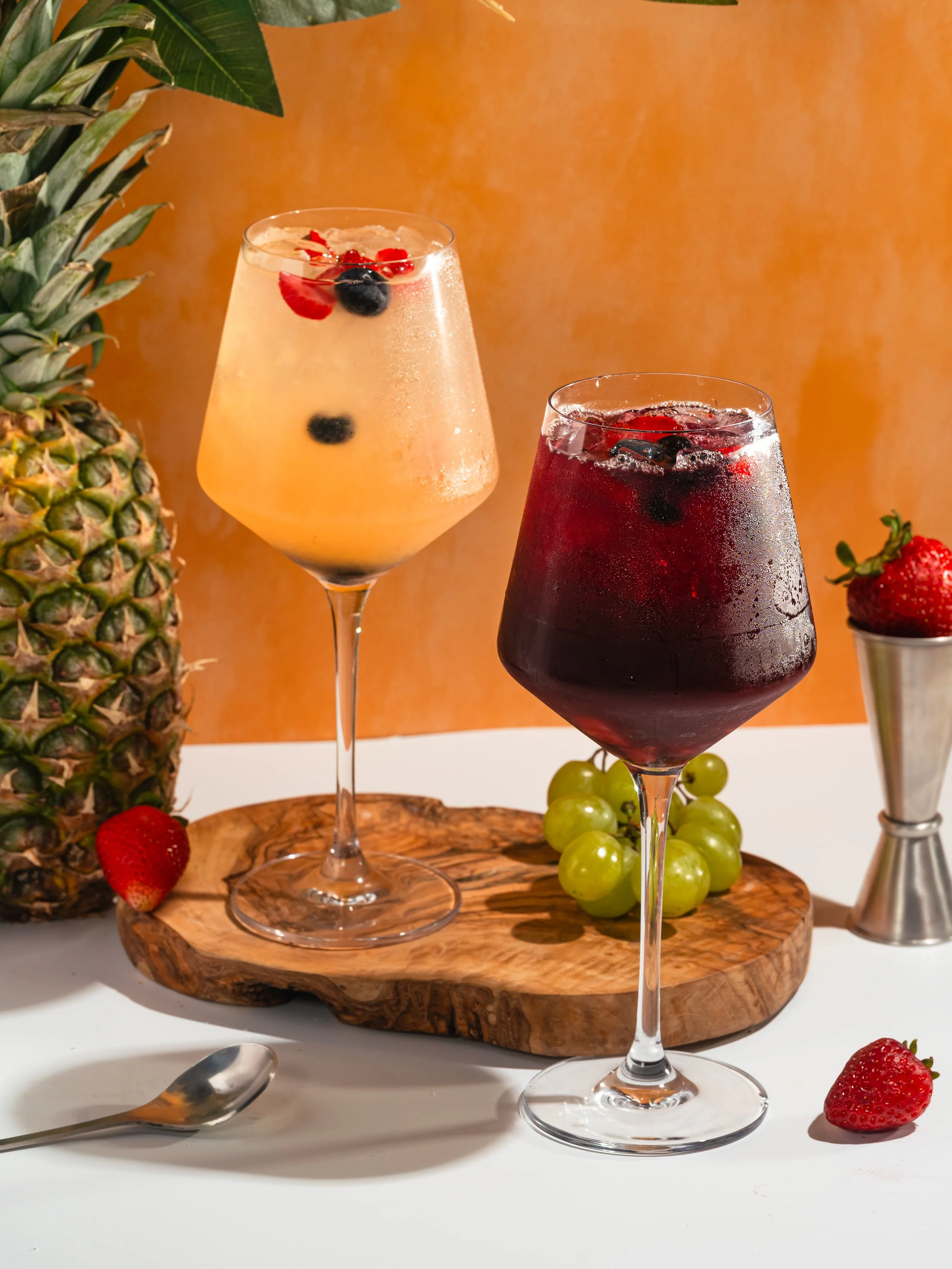 Two glasses of fruit drinks with berries, a pineapple on the left, and strawberries and grapes on a wooden tray, set against an orange background.
