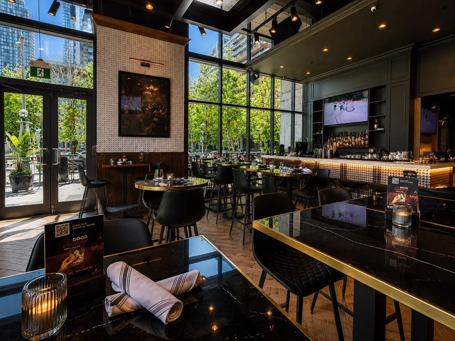 Interior of a modern restaurant with large glass windows, black tables with gold trim, and black chairs. The bar area is in the background with a television screen showing ice hockey. The restaurant has a mix of natural and artificial lighting, with potted plants outside visible through the windows.