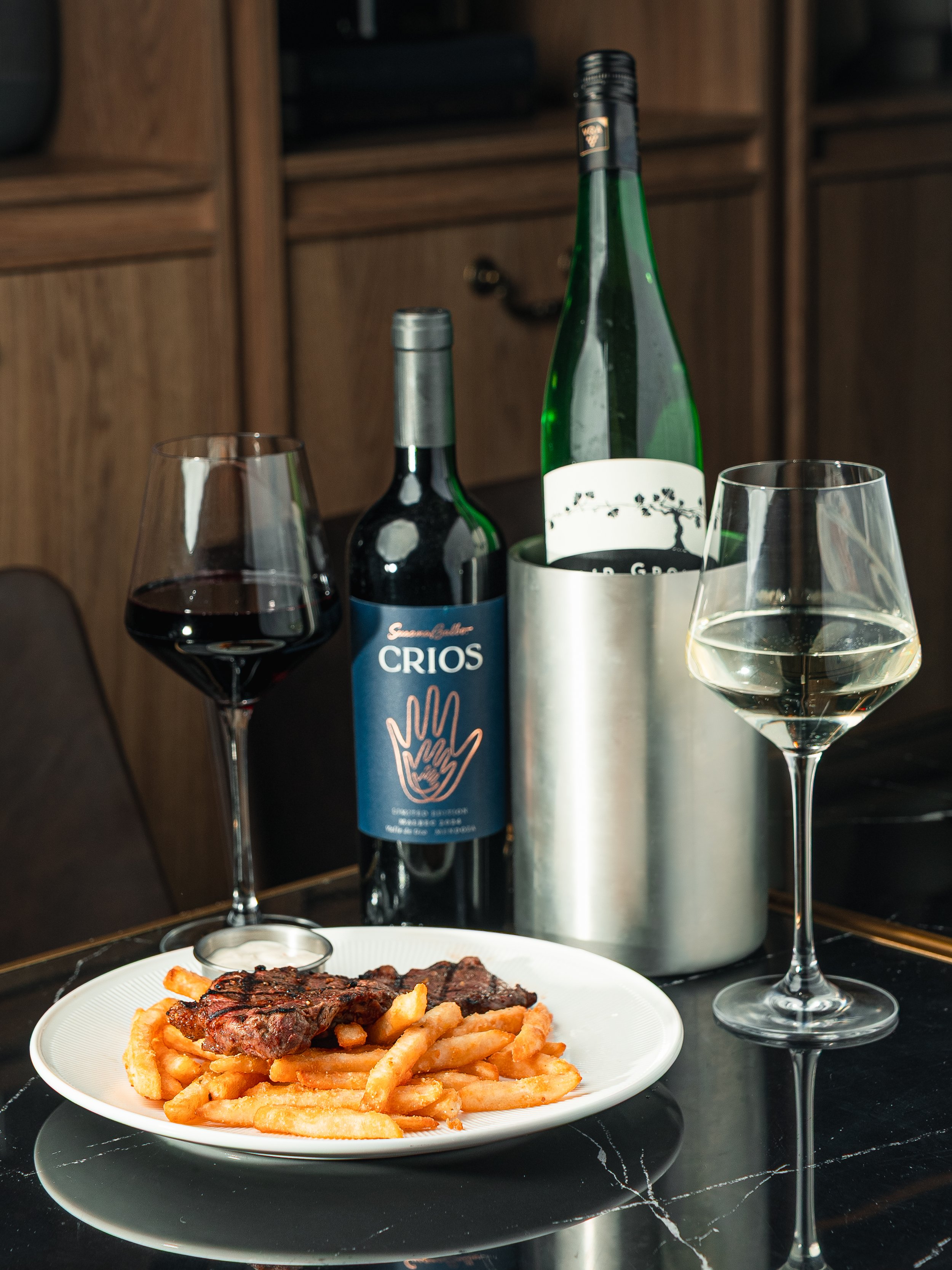 A plate of French fries topped with grilled meat, with a glass of red wine and a glass of white wine on a black marble table, with two bottles of wine in the background.