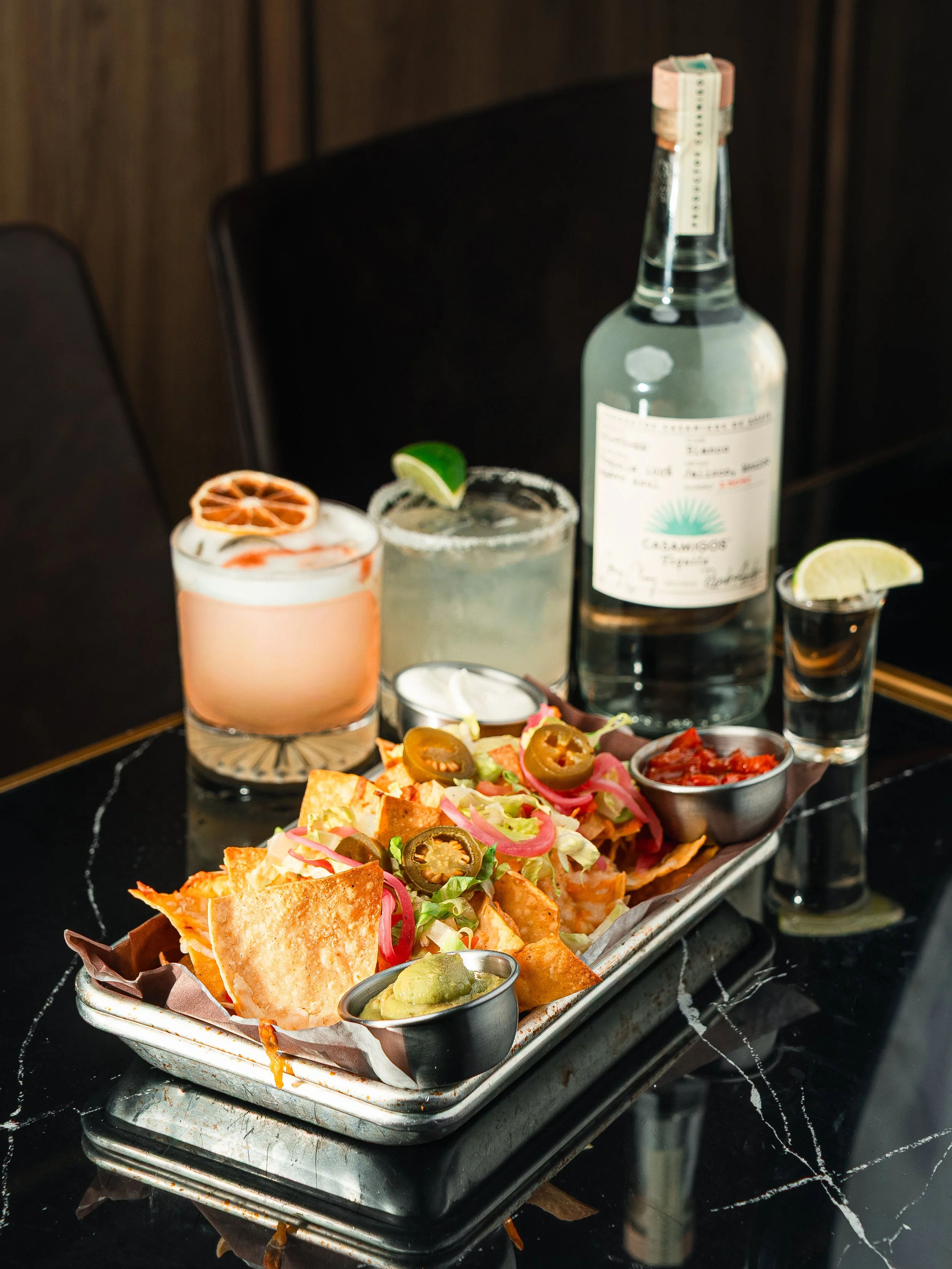 A tray of nachos topped with jalapenos, pink onions, and cheese, with bowls of guacamole and salsa, in front of a drinks and bottles on a black marble table.