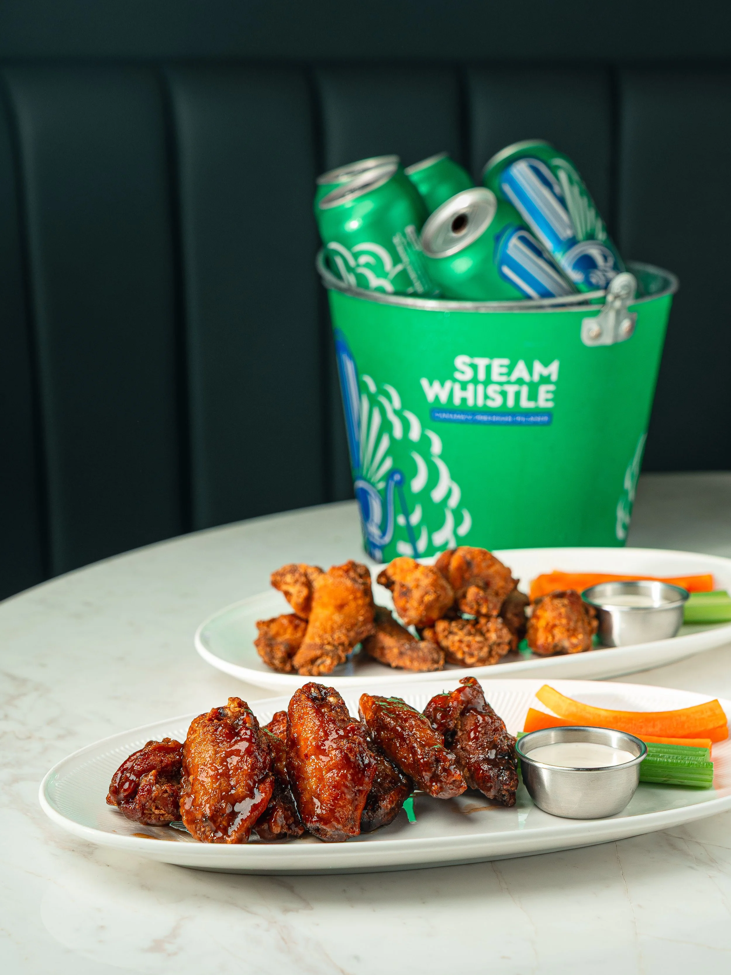 A plate of barbecue chicken wings with dipping sauce and vegetable sticks in front of a container of canned soda, with additional food plates visible in the background.