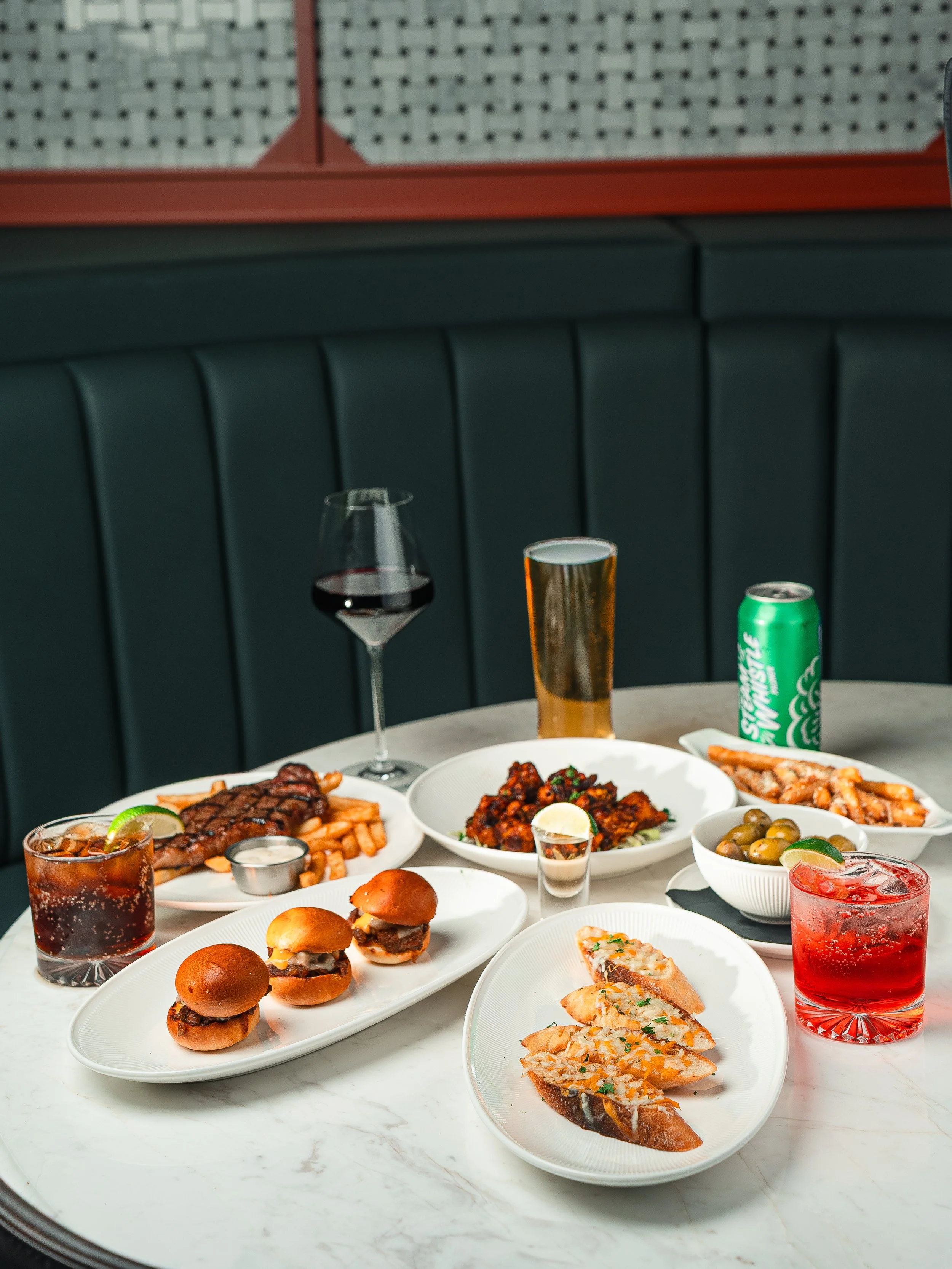 A table with various dishes including sliders, pasta, olives, and drinks like cola, cocktail, and soda.
