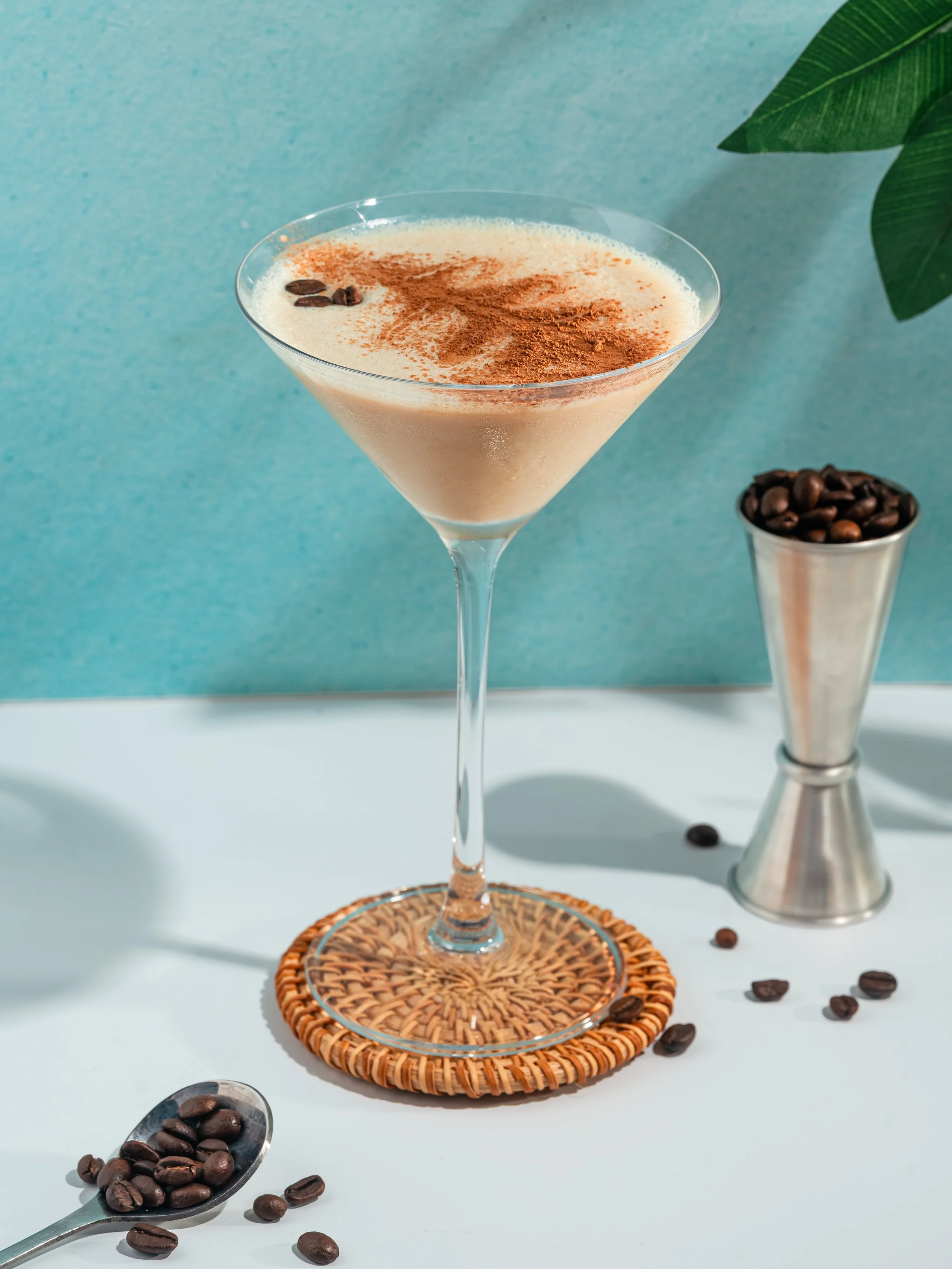 A glass of creamy coffee-flavored cocktail garnished with cocoa powder and coffee beans, with a spoon and scattered coffee beans on a white surface and a blue background.