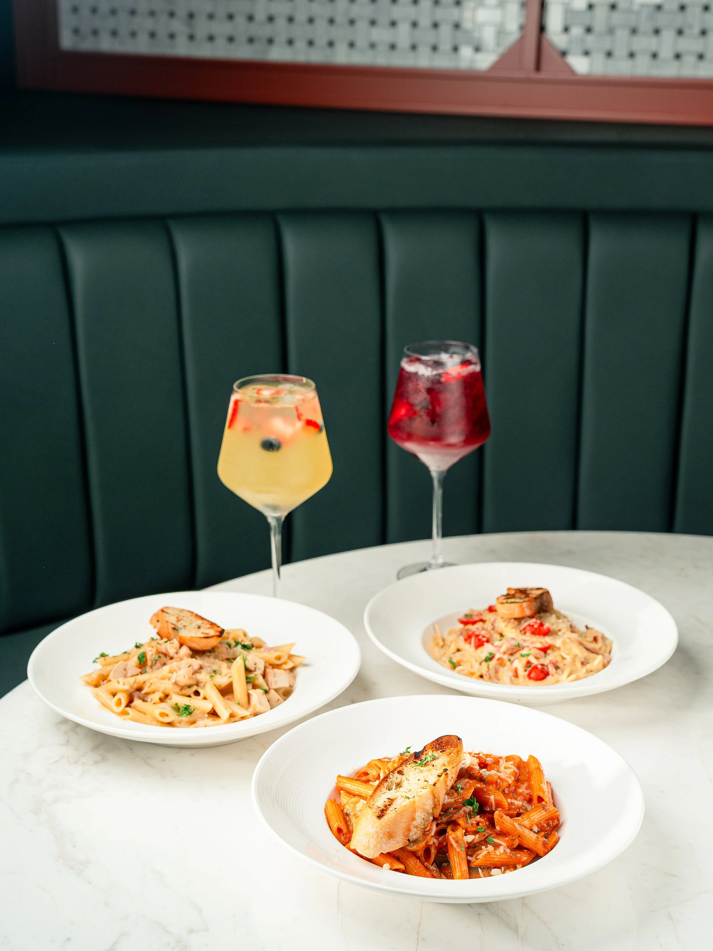 Three bowls of pasta with different sauces and a piece of garlic bread on a white marble table, with two glasses of colorful beverages behind them.