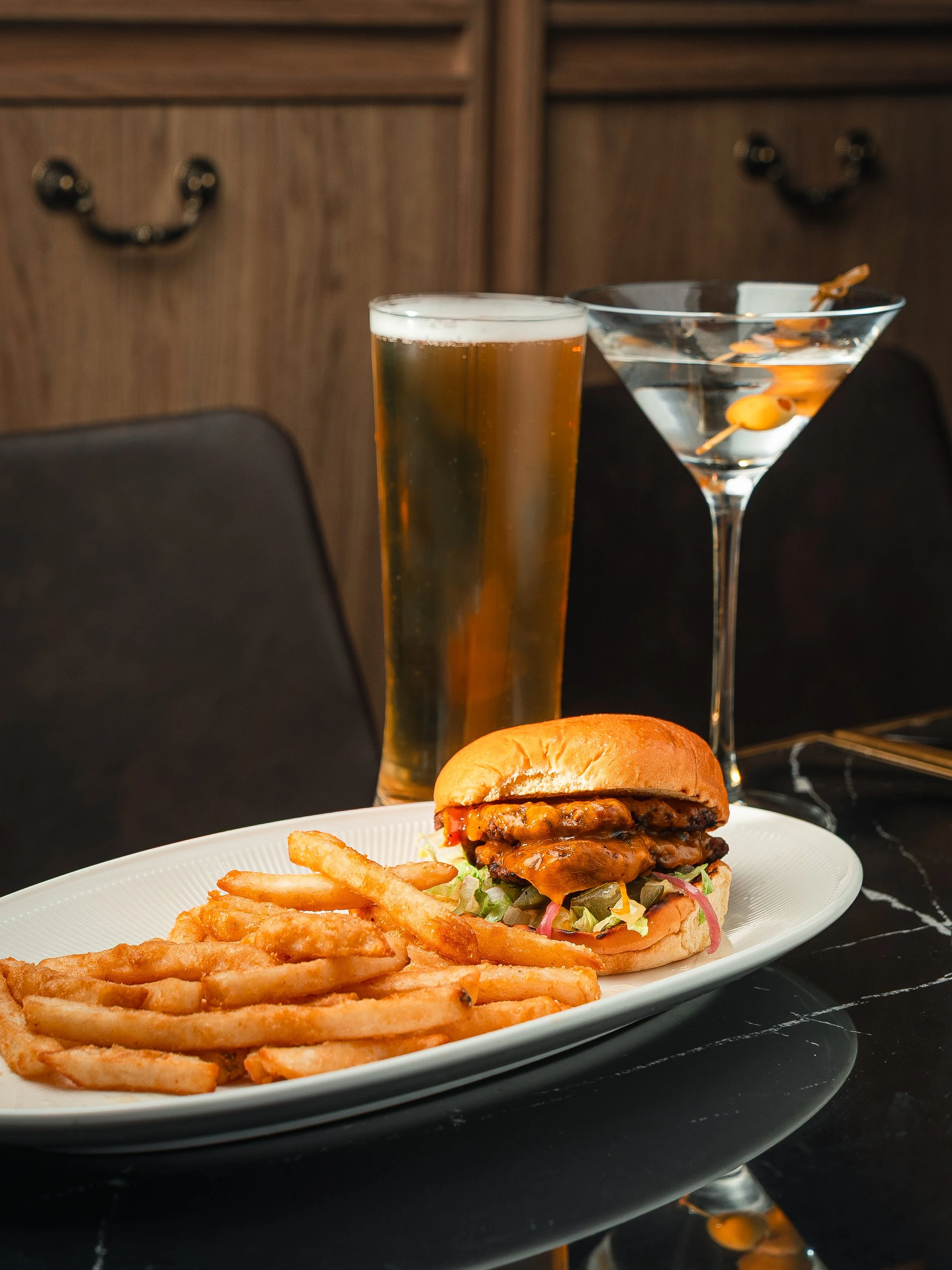 A plate of French fries, a cheeseburger with lettuce, pickles, onions, tomato, and sauce, a pint of beer, and a martini with olives on a toothpick on a black table.