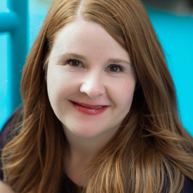 Close-up of a smiling woman with red hair and fair skin, outdoors with a blue background.