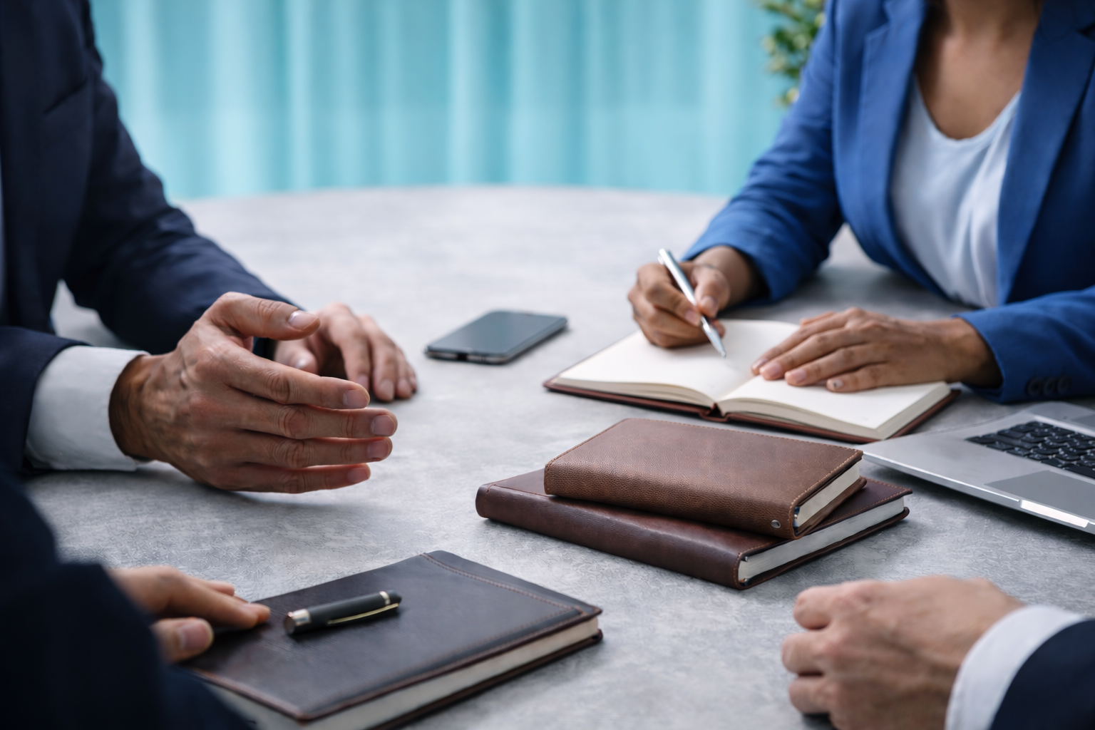 People's hands and arms at a business meeting table, with notebooks, a pen, a smartphone, and a laptop visible.
