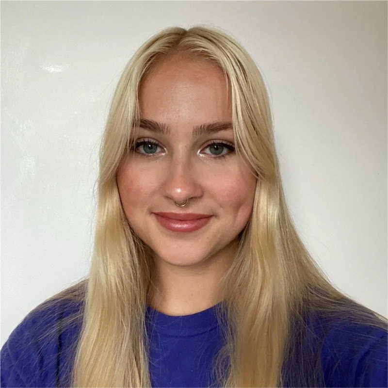 Close-up of a young woman with long blonde hair, blue eyes, and a septum piercing, wearing a purple shirt, smiling softly against a plain white wall.